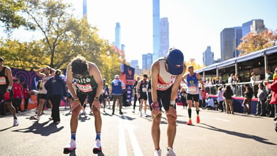 Two runners bent over with their hands on their knees with the TCS New York City Marathon finish line and other runners behind them