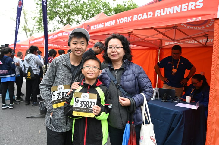 Two children with their race bibs standing with their mom in front of a Rising New York Road Runners tent
