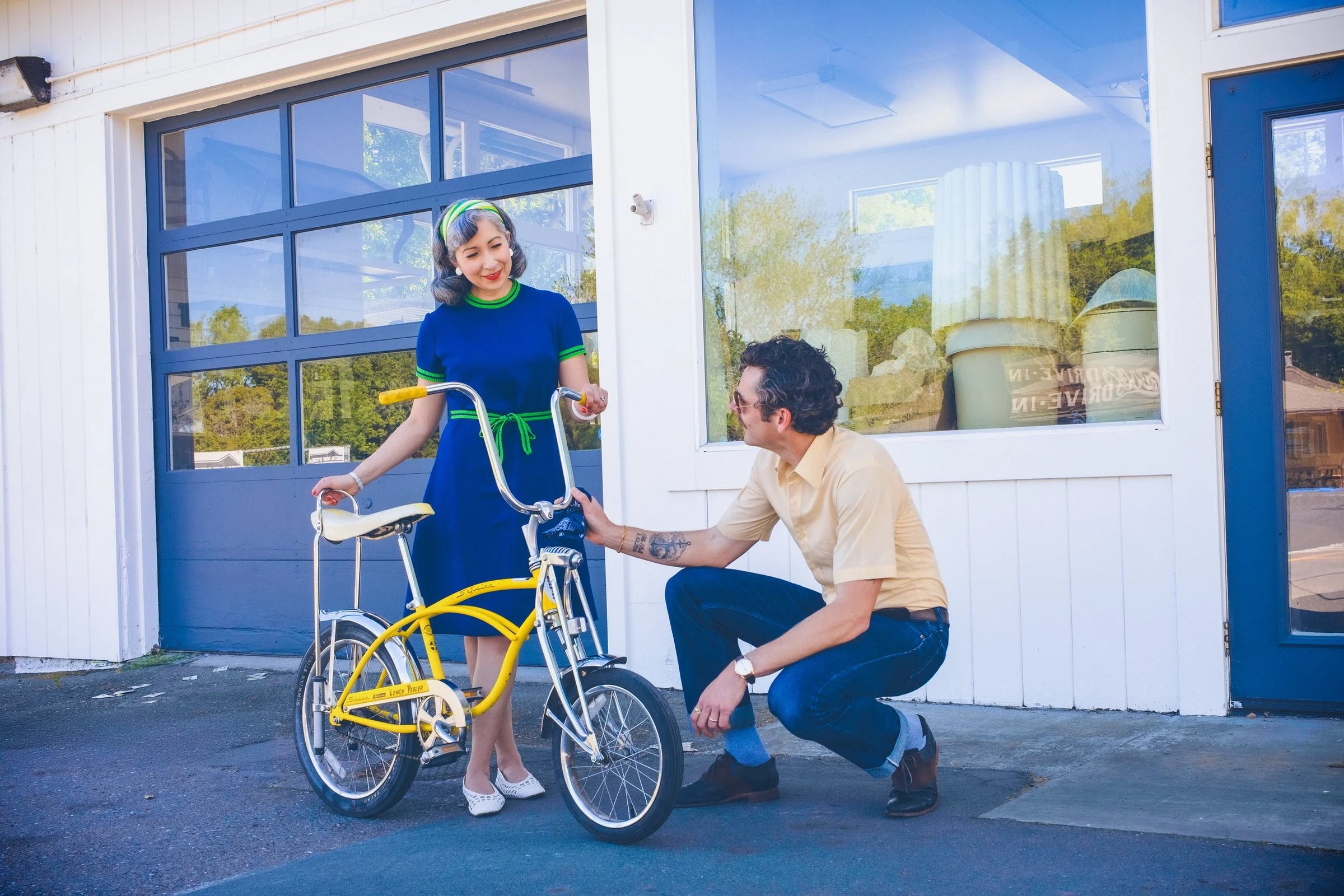 A woman in a blue dress with green accents stands next to a yellow bicycle while a man in a beige shirt kneels beside her, both smiling in a vintage-style setting.