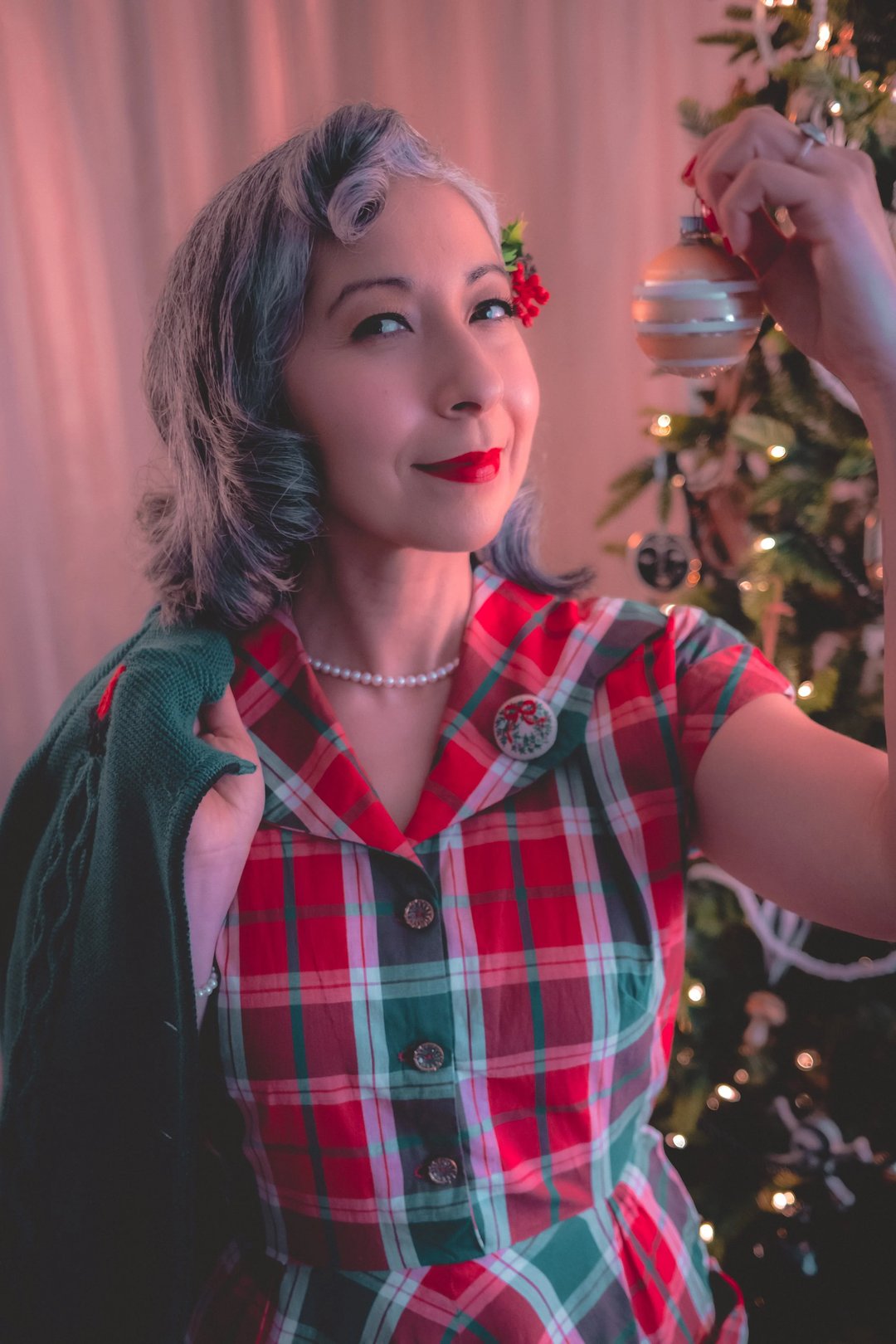 Woman with short, wavy gray hair wearing a red and green plaid dress holds a Christmas ornament in front of a decorated tree.