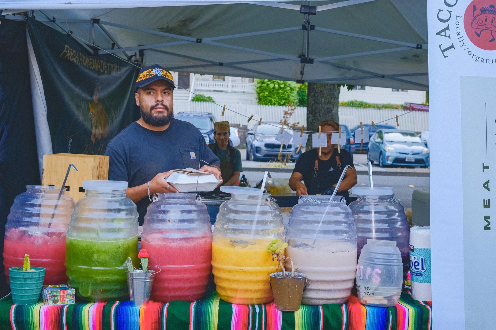 A vendor at a market stand holds a plate while colorful beverage dispensers sit in front of him.