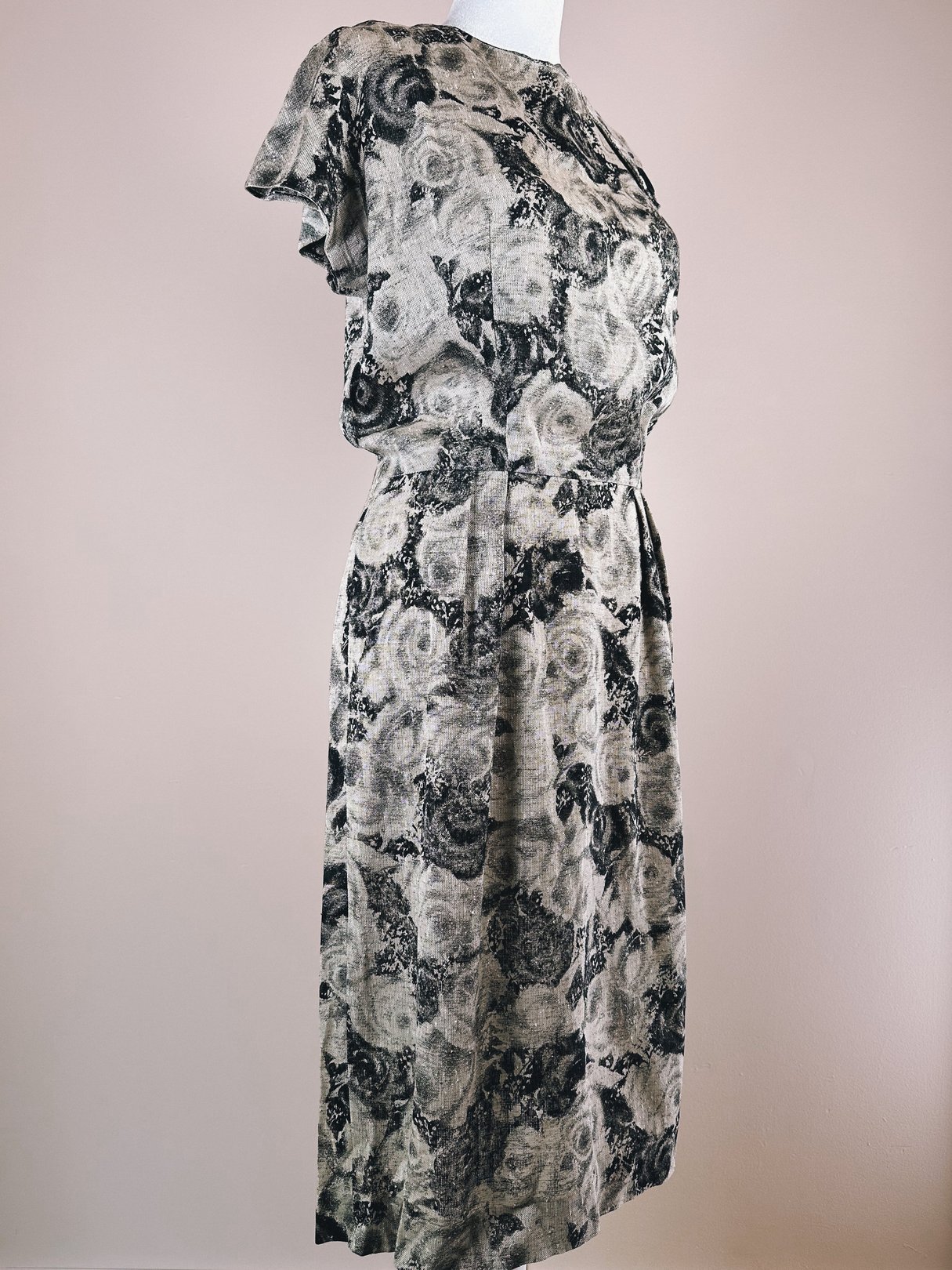 A 1950s women's dress featuring a black and cream rose pattern, with short flutter sleeves and a fitted waist, displayed on a mannequin against a light background.