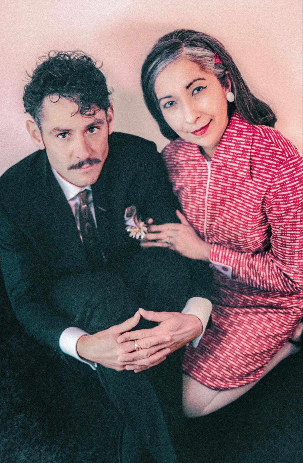 A man in a suit and a woman in a patterned dress pose together with a stylish, vintage-inspired aesthetic.