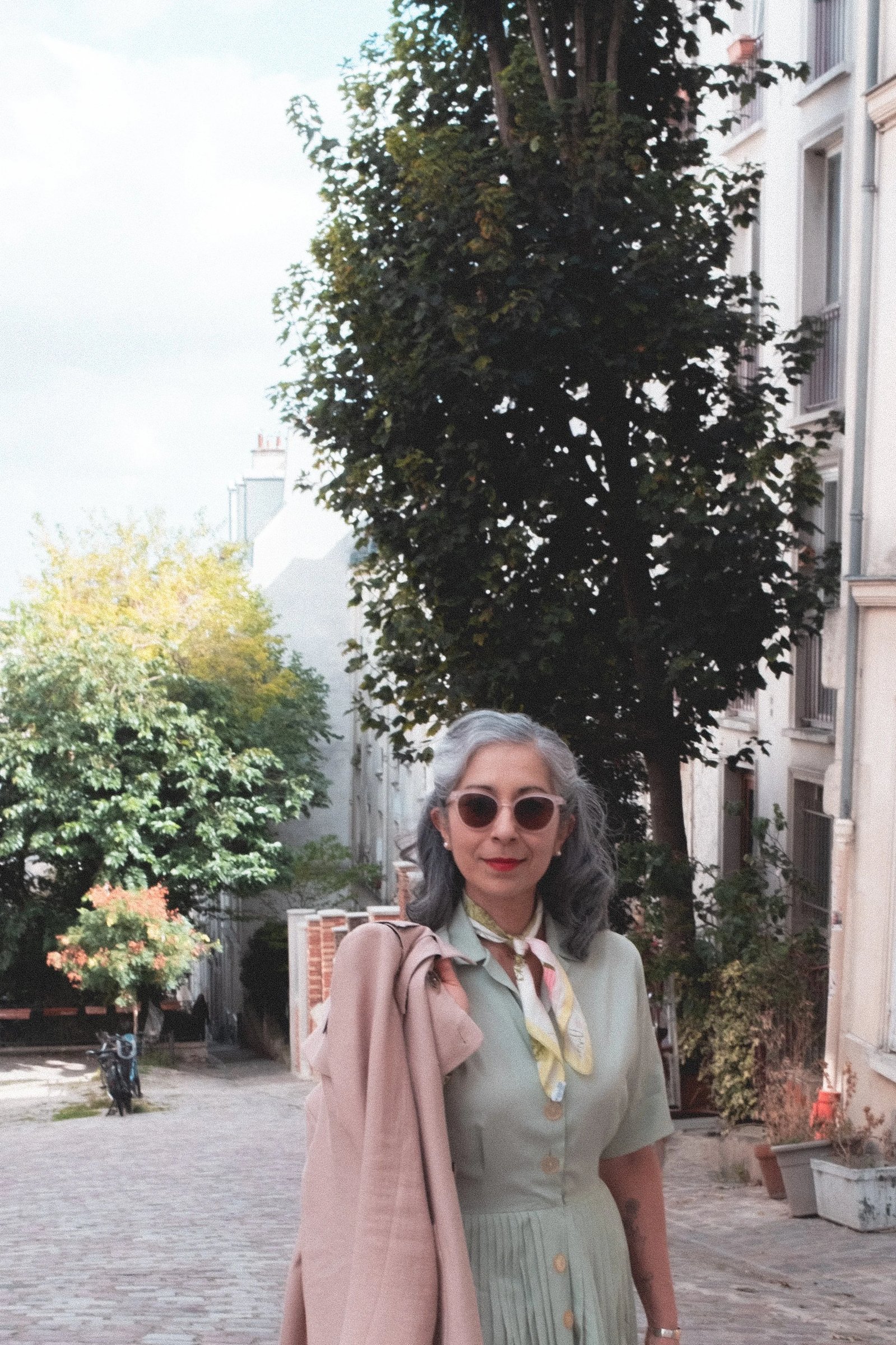 A woman with silver hair wearing a mint green dress and sunglasses stands on a cobblestone street lined with trees and buildings.