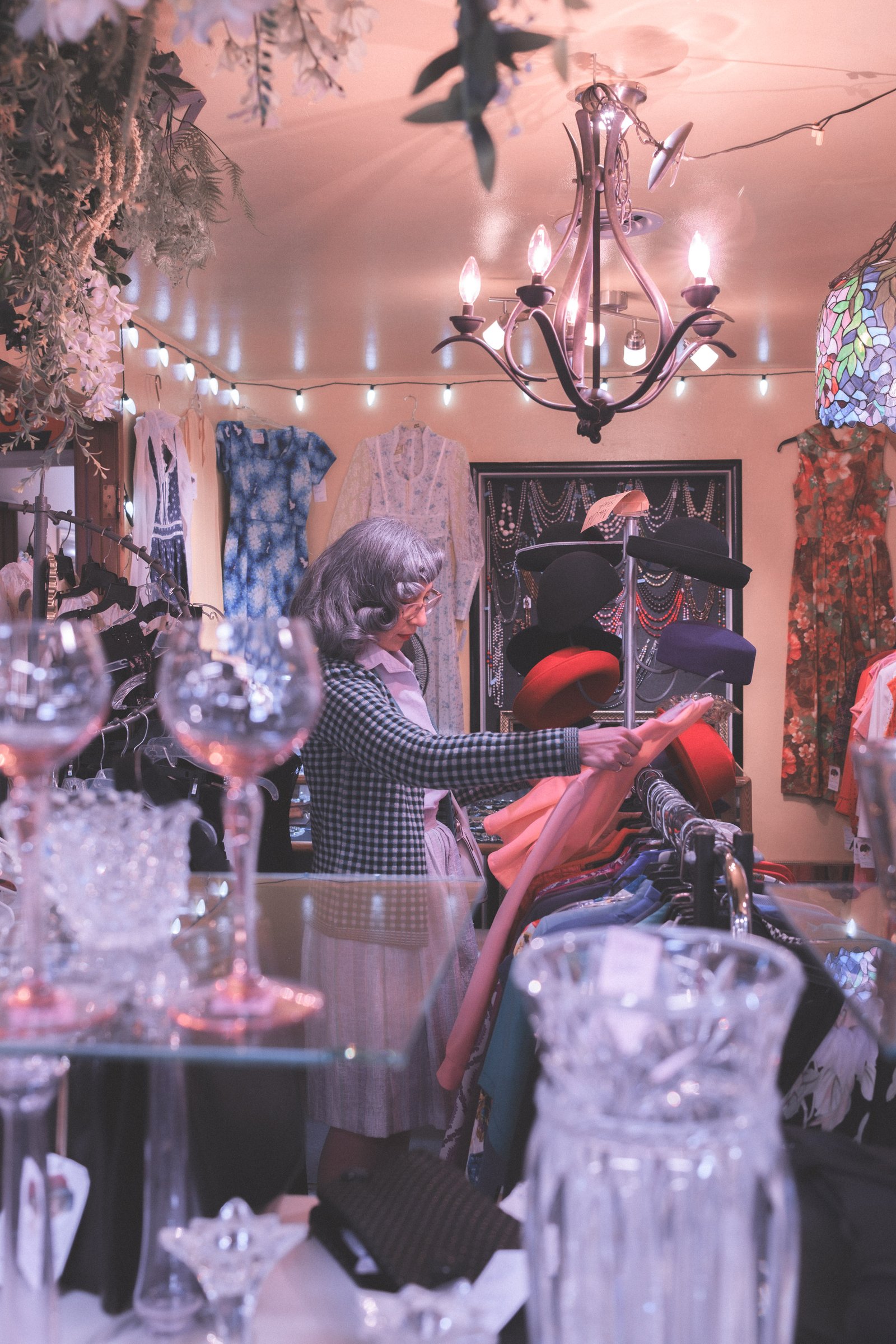 Sheet Music Stories gallery image: A woman in a vintage store examines colorful clothing displayed on a rack, surrounded by various decorative items and soft lighting.