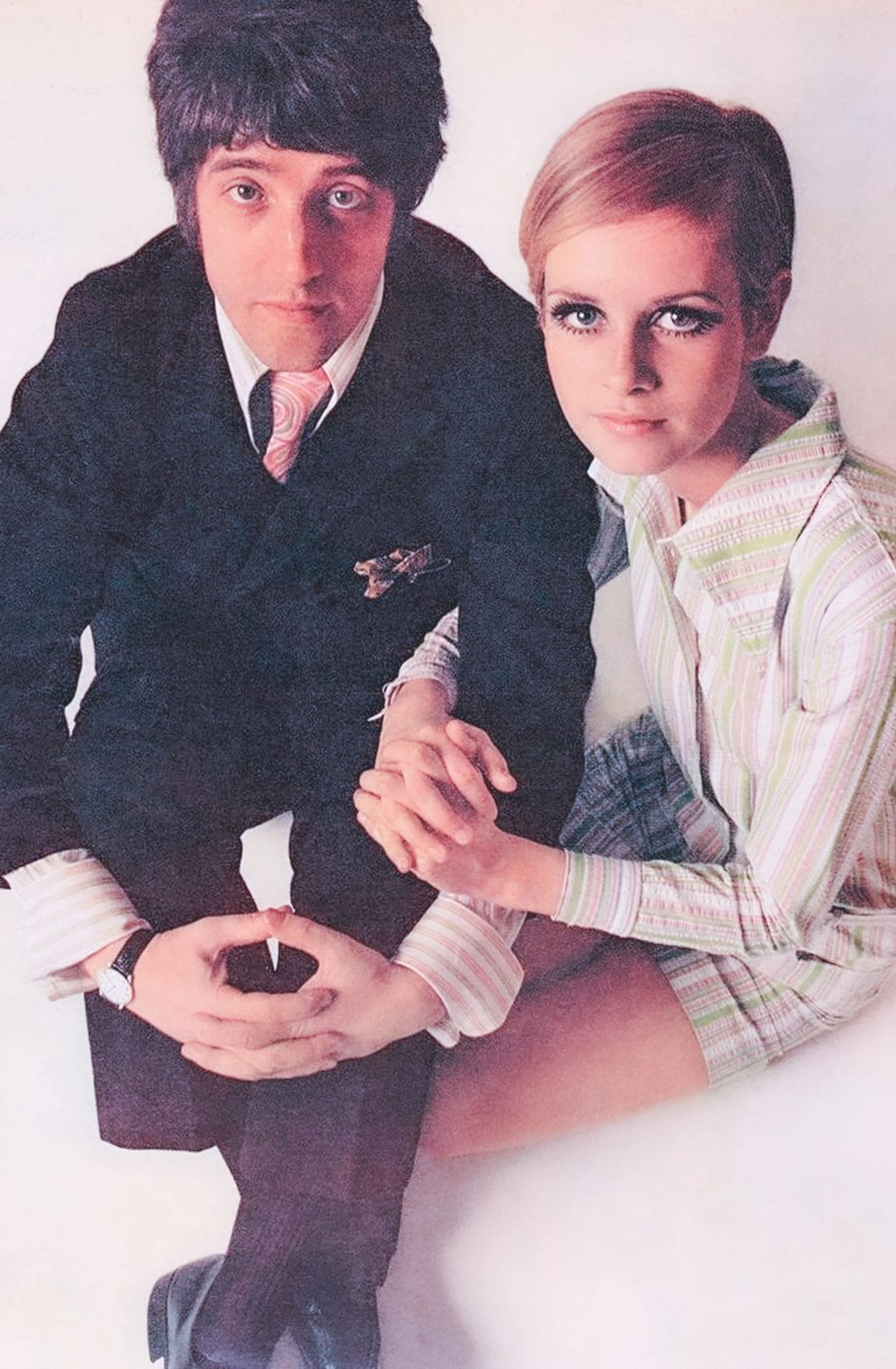 A young man in a suit and a young woman in a striped dress sit closely together, holding hands against a soft, neutral background.