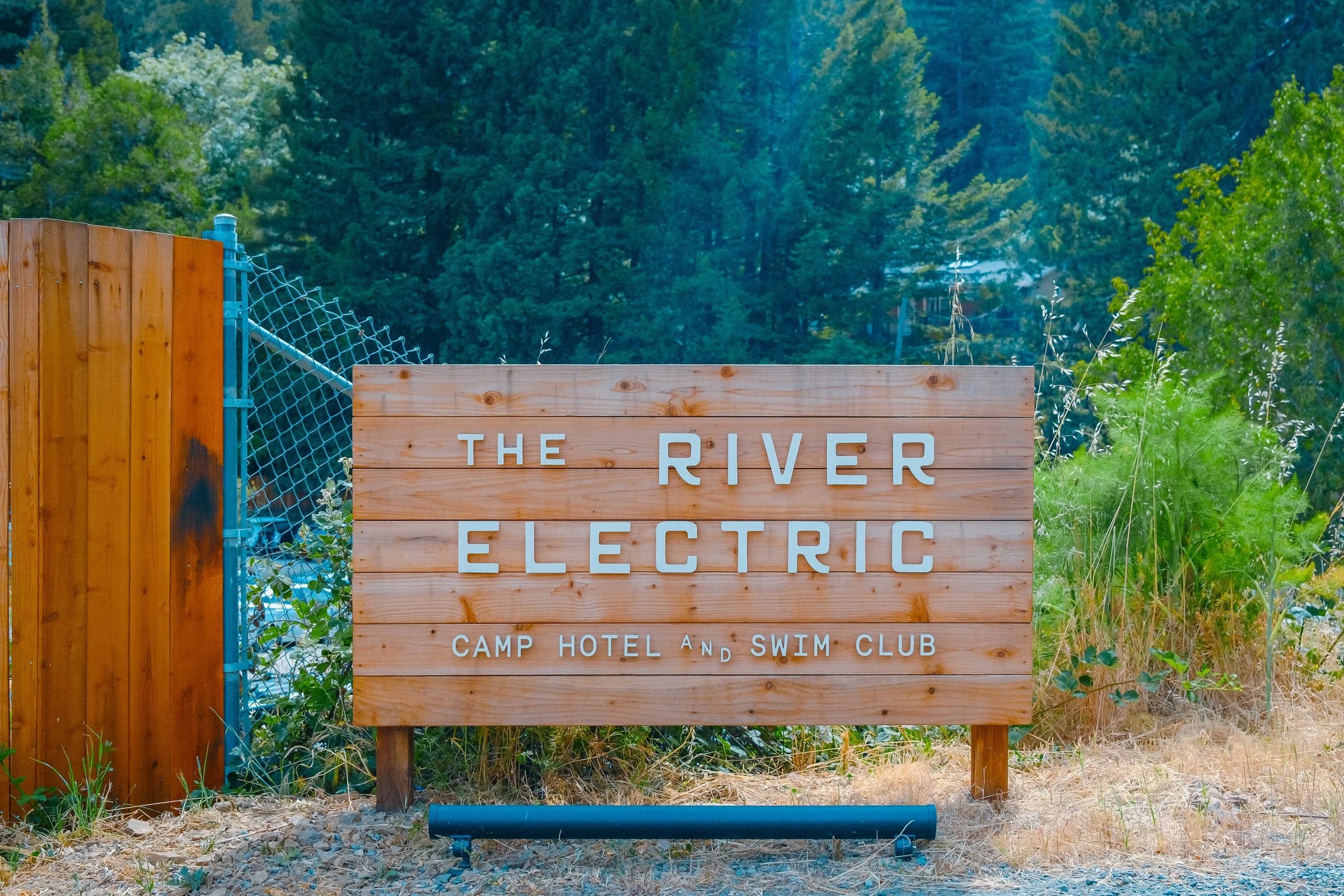 Wooden sign for The River Electric Camp Hotel and Swim Club surrounded by greenery.