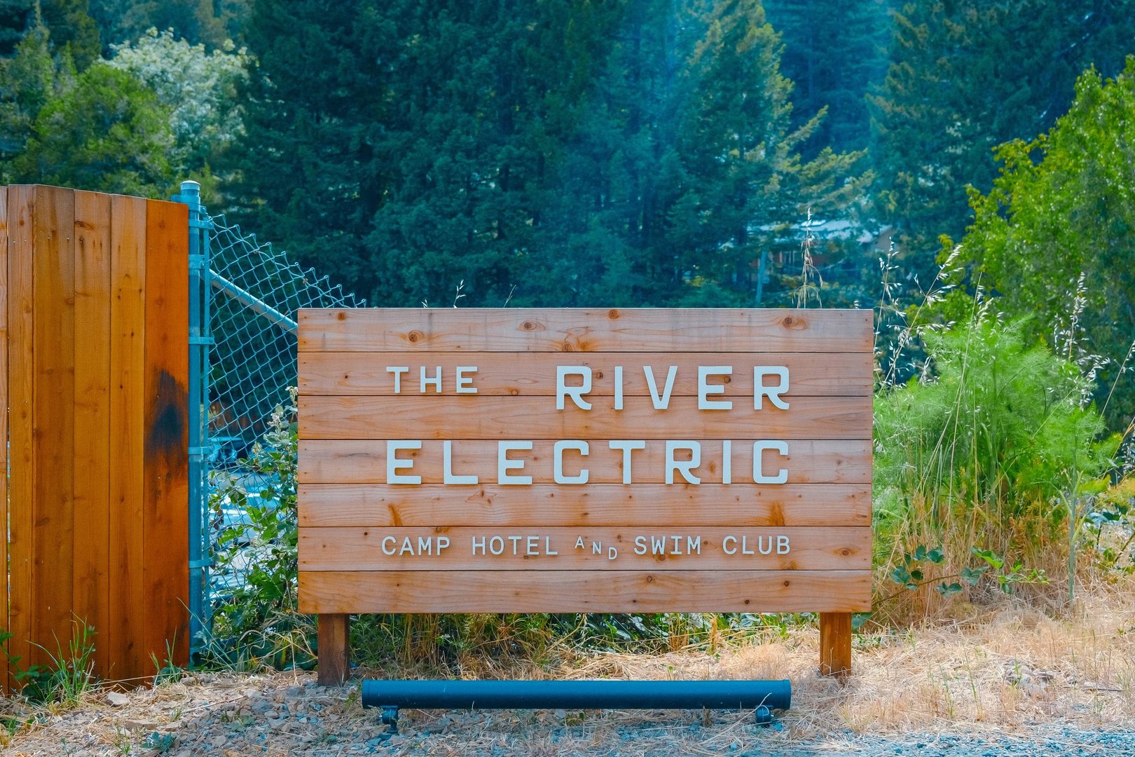Wooden sign for The River Electric Camp Hotel and Swim Club surrounded by greenery.
