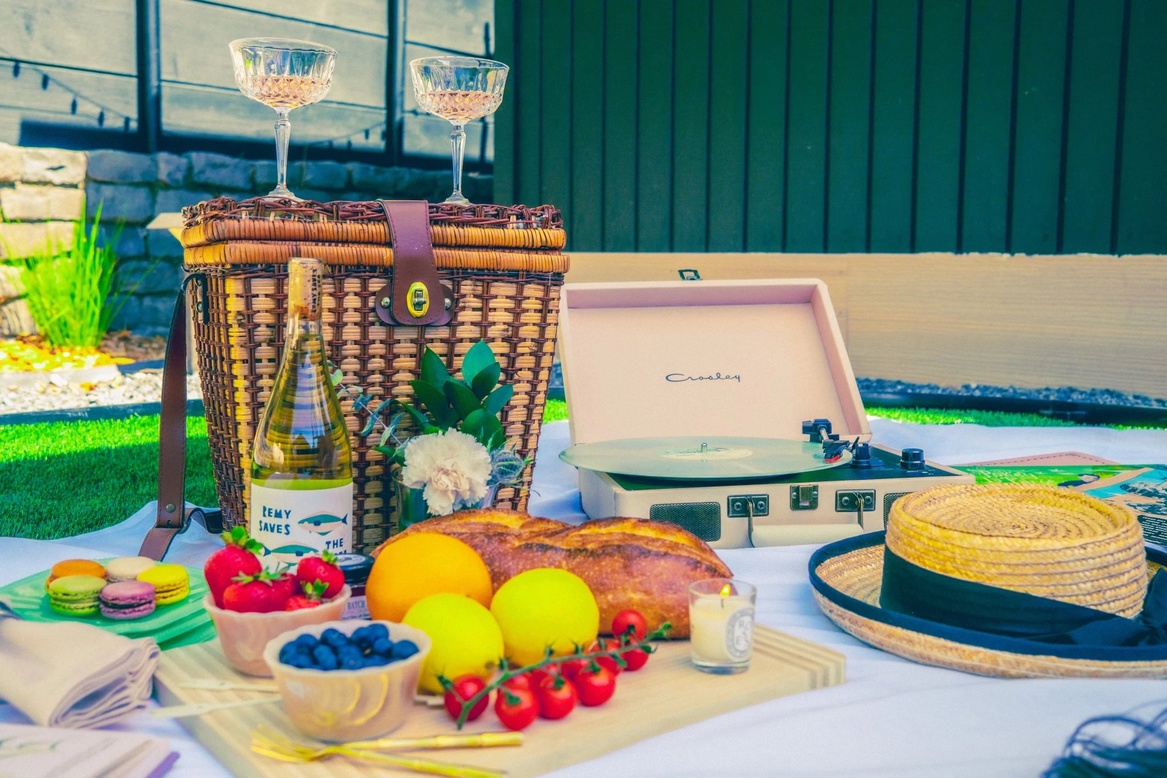A picnic setup featuring a wicker basket, wine, fresh fruits, pastries, a record player, and glasses on a blanket.