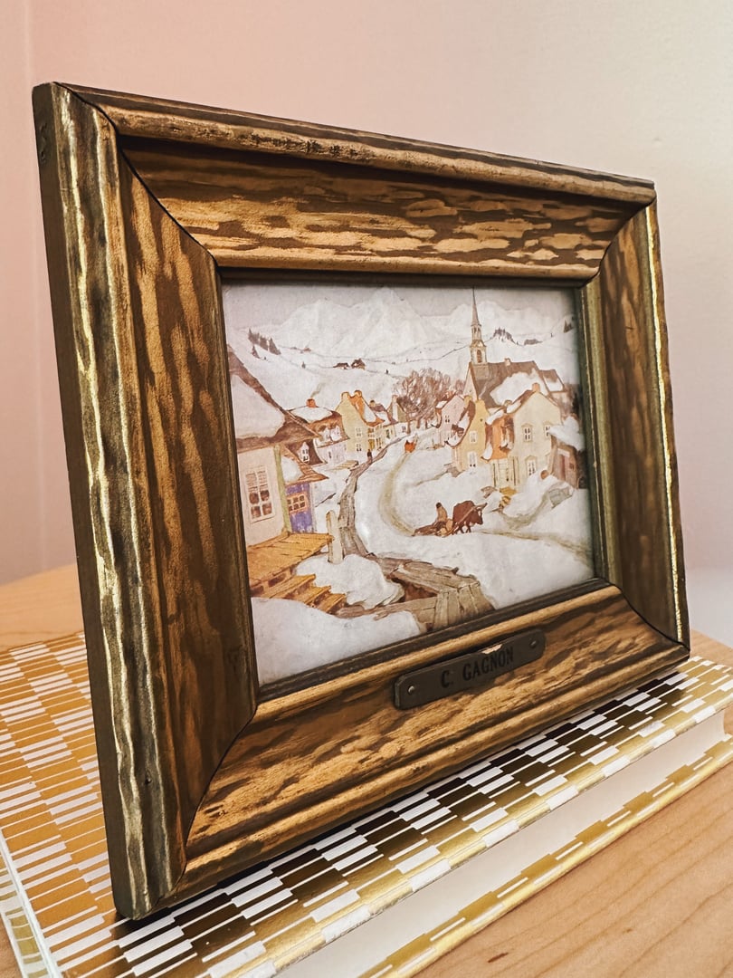Framed art piece titled "Village in the Laurentian Mountains" by C. Gagnon, featuring a winter landscape with a church, in a gold and brown wooden frame, displayed on a wooden surf