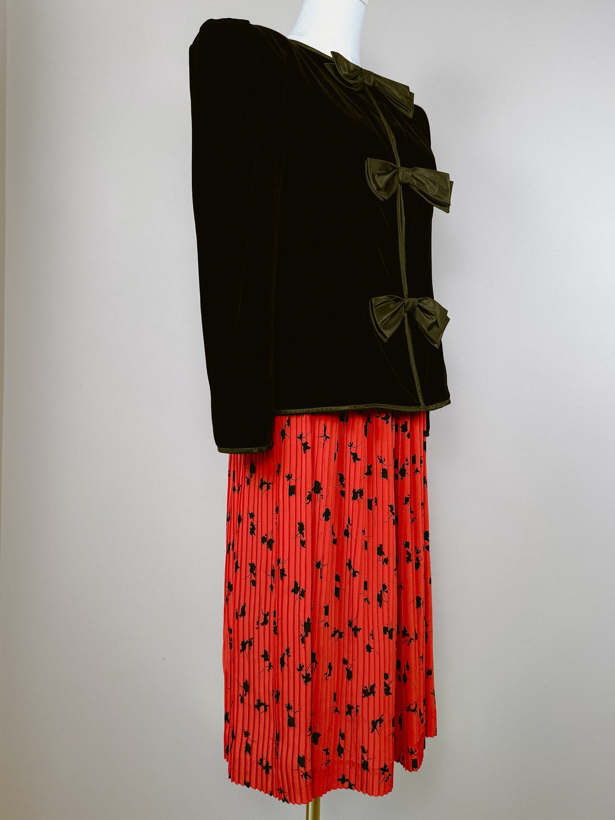 Black velvet 1980s women's jacket featuring three bows, styled over a red pleated skirt with a floral pattern.