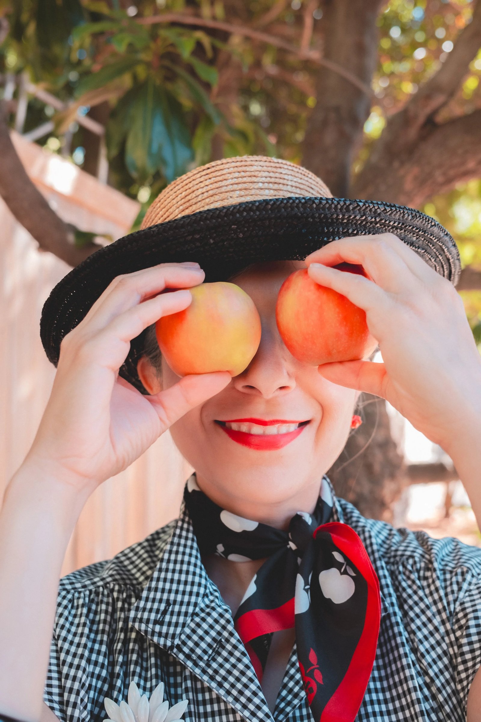 A woman in a black and white checkered outfit and straw hat holds two apples in front of her face, smiling.