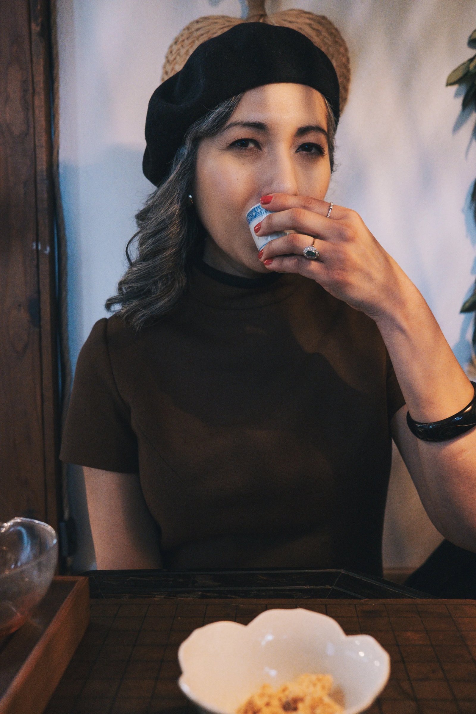 A woman in a beret sips from a small cup while seated at a table with a bowl of food in front of her.