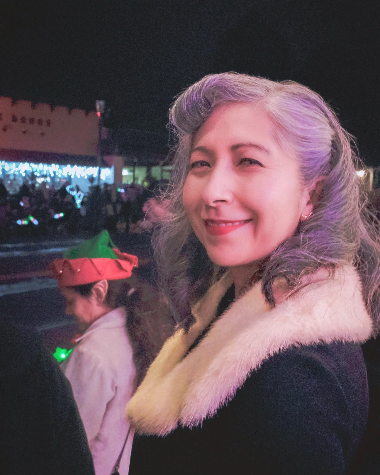 A woman with silver hair and a fur collar smiles at a festive evening event.