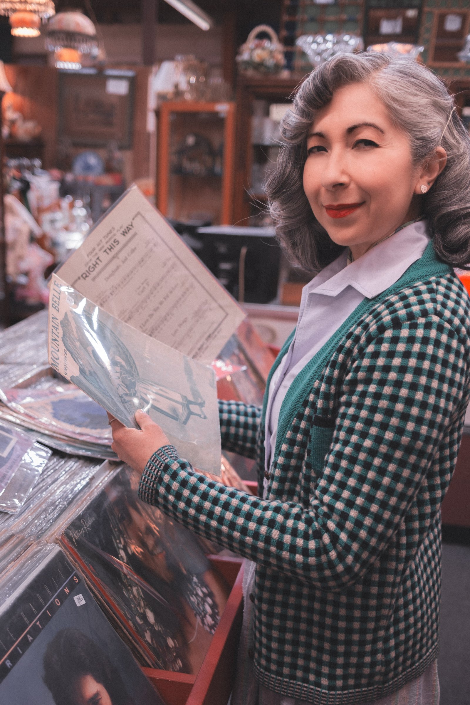 Sheet Music Stories gallery image: A woman in a checkered cardigan holds a sheet of music while standing in an antique shop surrounded by vinyl records.