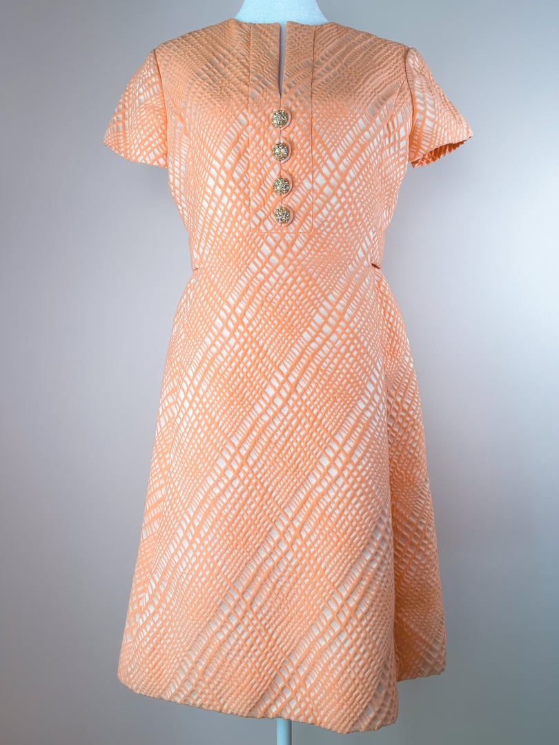 The Orange Jubilee Dress features a textured pattern in shades of orange, short flutter sleeves, and a buttoned front design typical of 1960s-style dresses.