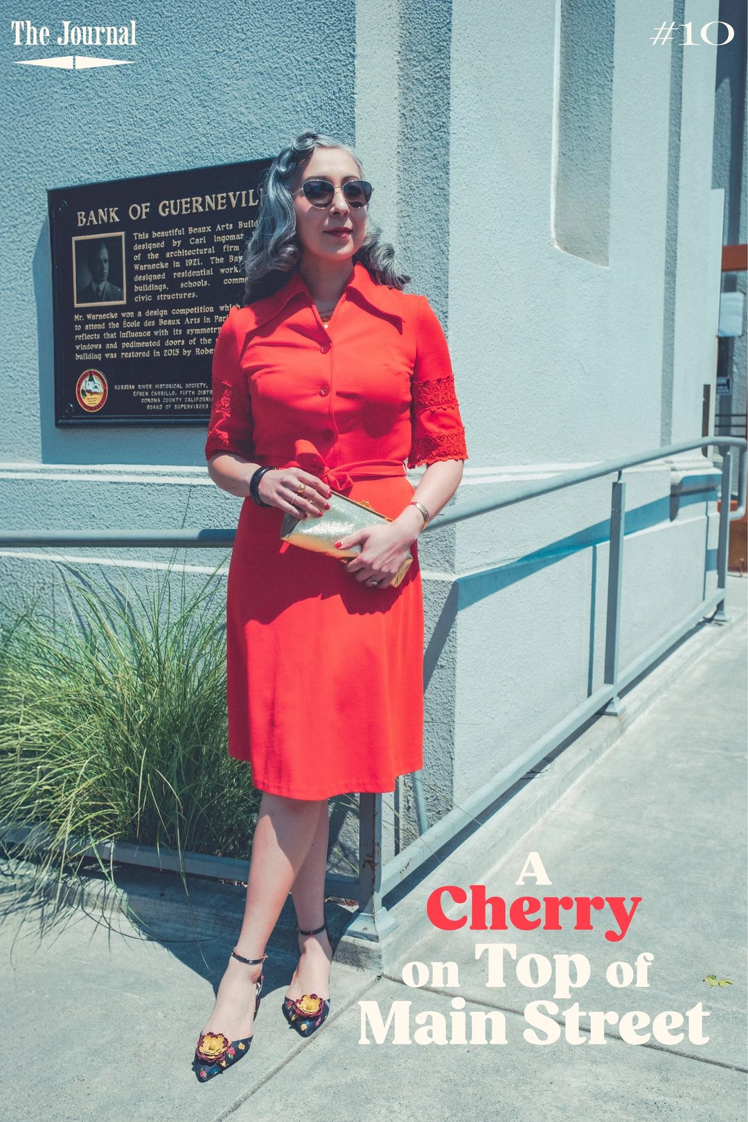 A woman in a red dress and sunglasses stands outside a building, holding a clutch purse.
