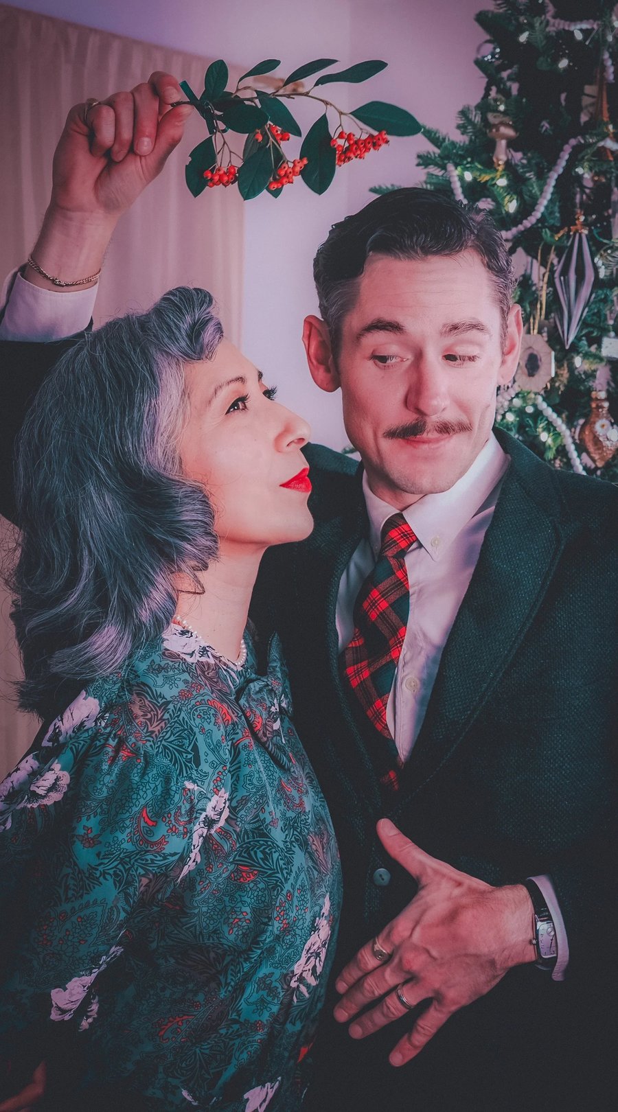 A man in a suit and tie gazes at a woman with gray hair and red lipstick, both dressed in festive attire against a holiday backdrop.