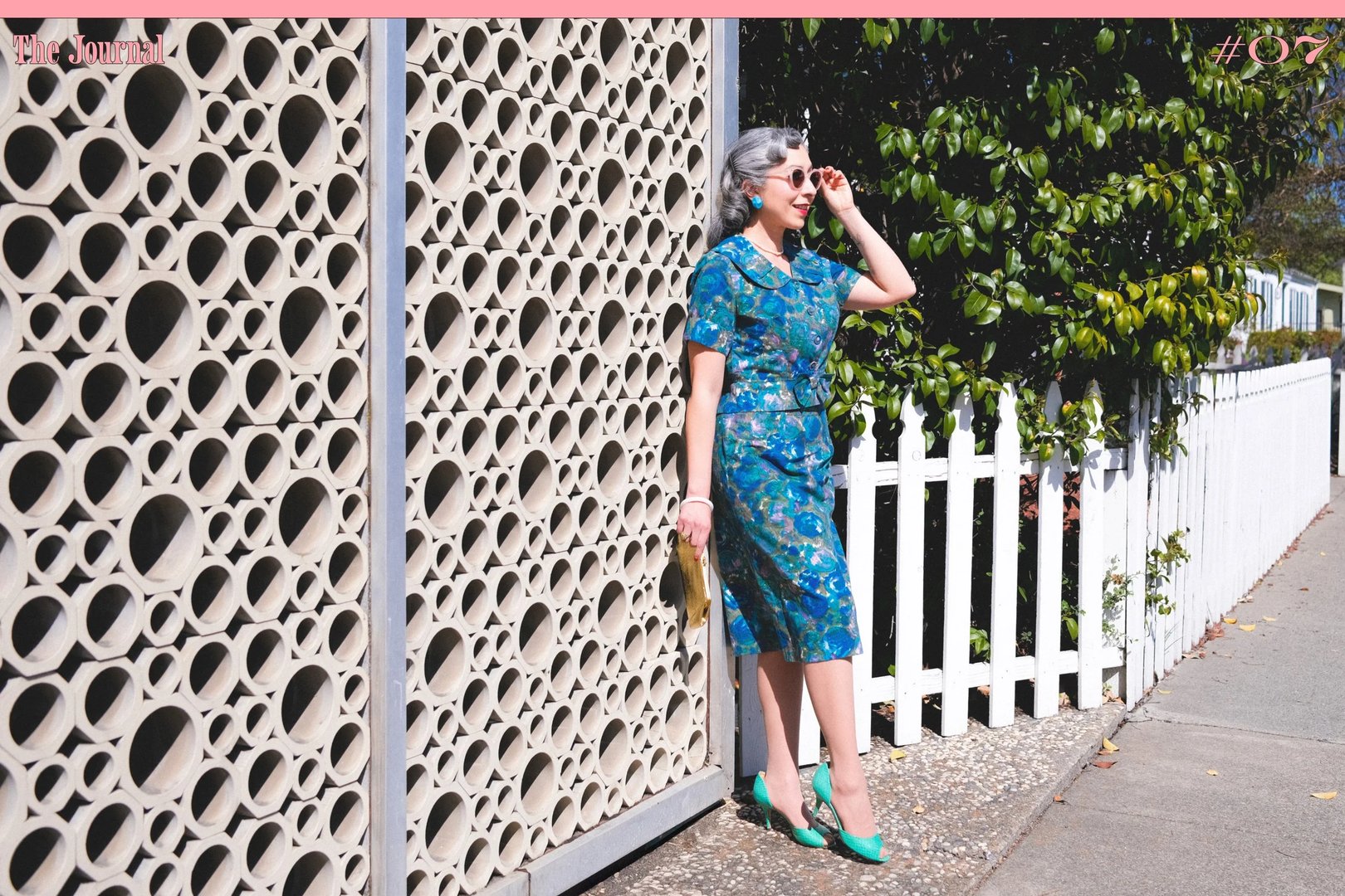 A woman in a turquoise floral dress and green shoes stands next to a textured wall, holding a drink.