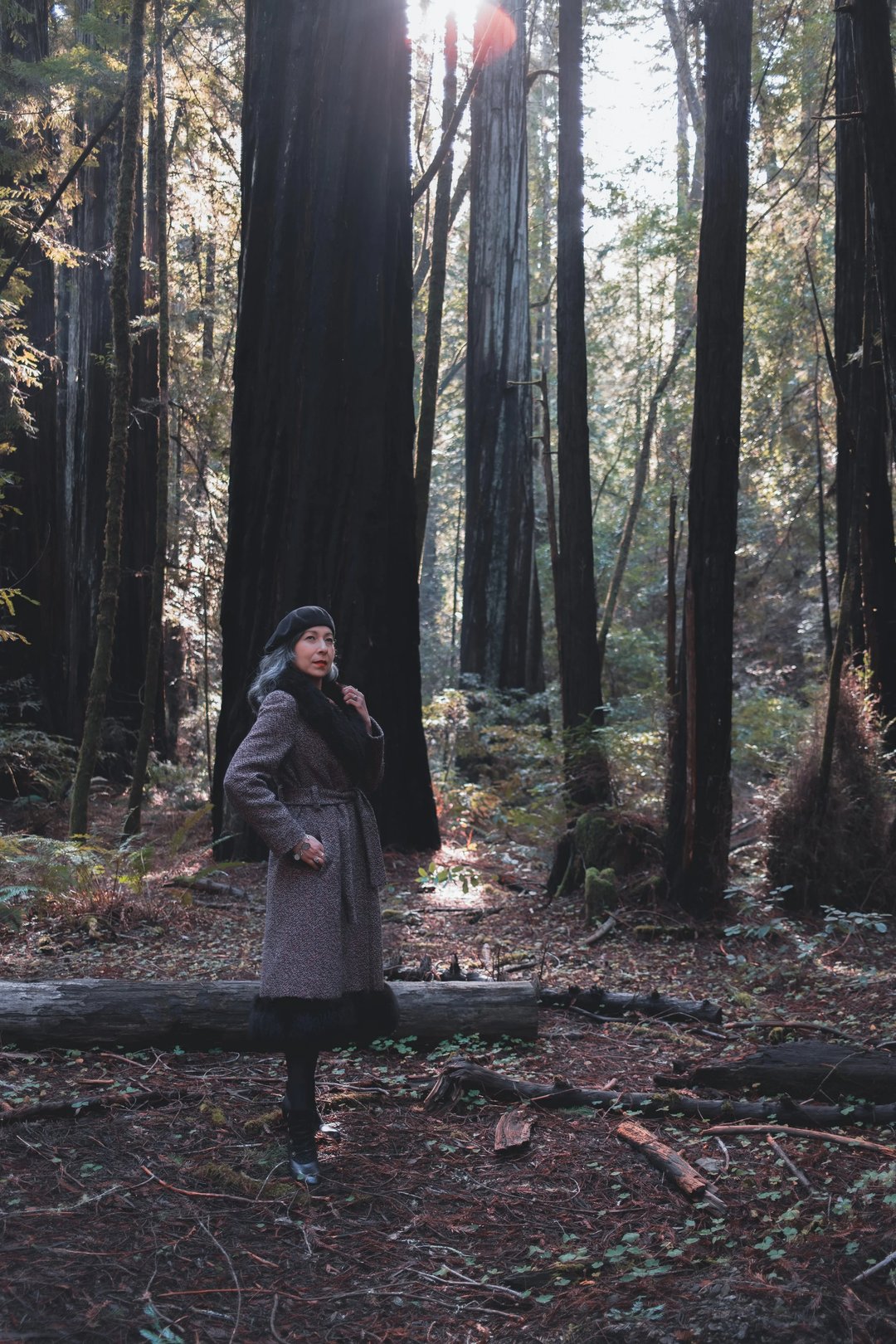 A woman stands in a forest of tall trees, wearing a long coat and looking off to the side.