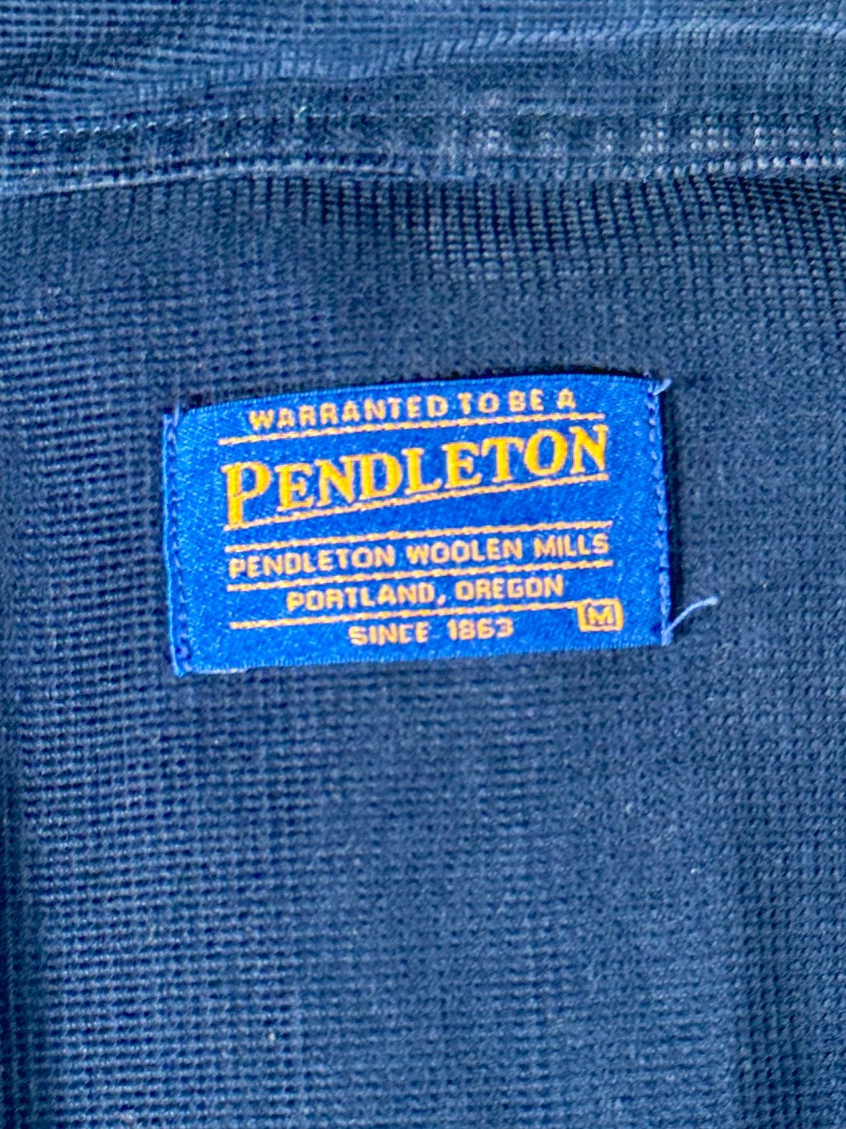 Navy blue tag displaying "Pendleton Woolen Mills" with the text "Warranted to be a Pendleton" and indicating a size medium.