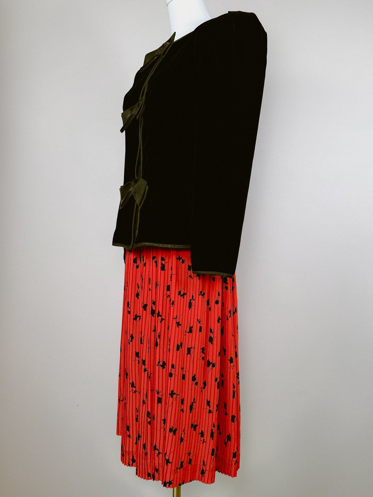 Black velvet jacket with a front bow detail, paired with a red pleated skirt featuring a floral pattern, styled on a mannequin.