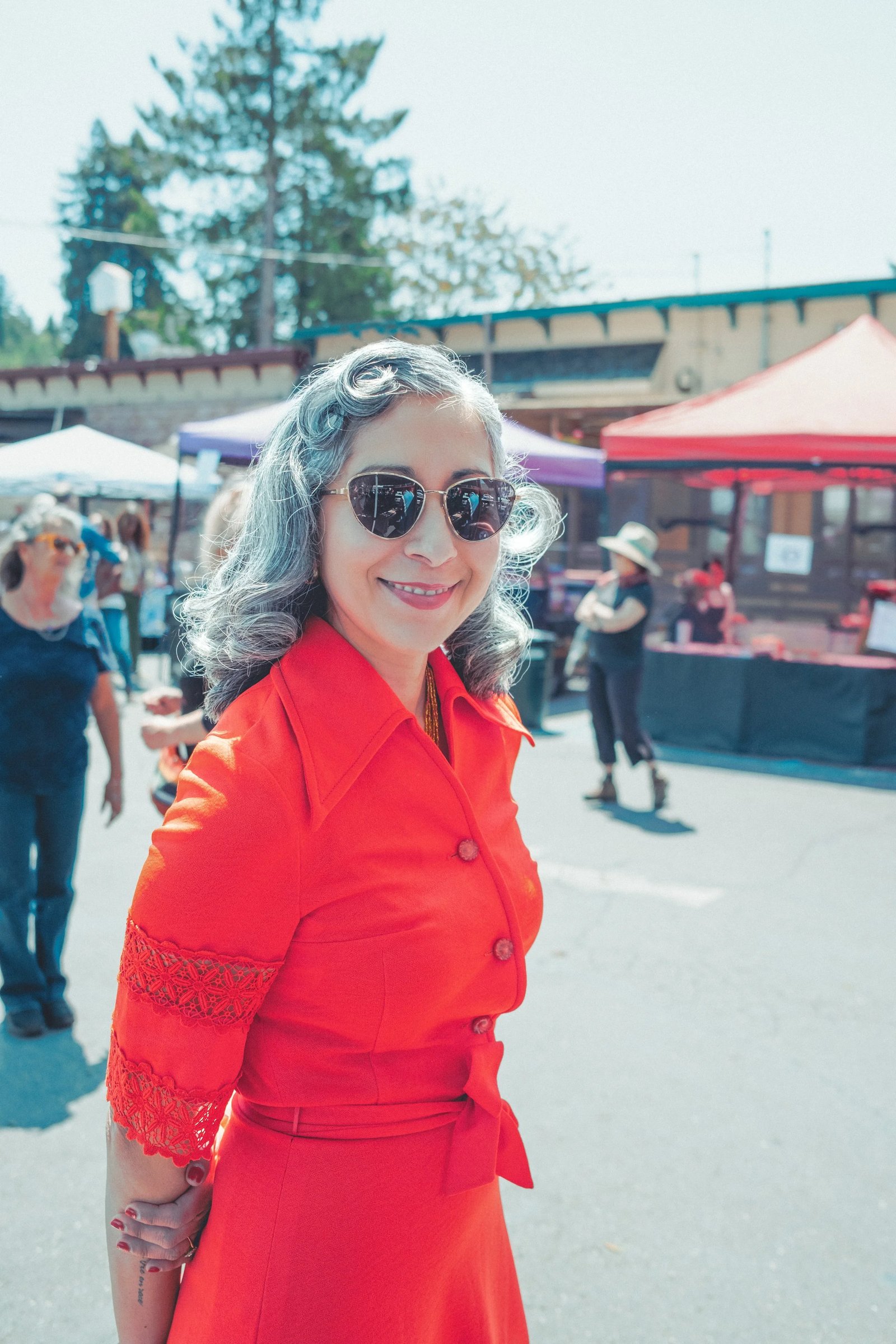A woman wearing a bright red dress and sunglasses smiles at the camera in a bustling outdoor market setting.