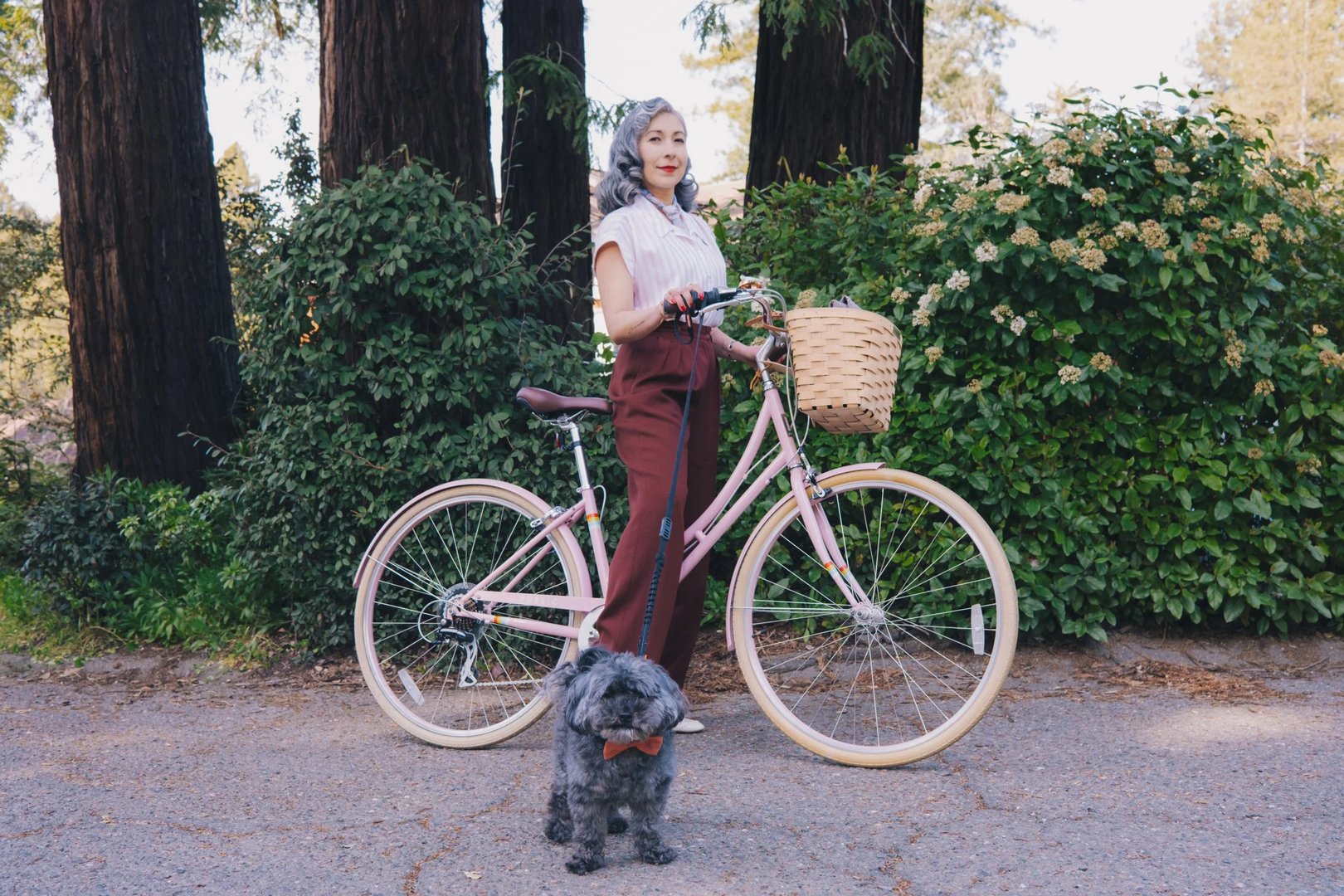 A woman stands beside a pink bicycle with a basket, holding the handlebars and accompanied by a small dog on a pathway surrounded by greenery.