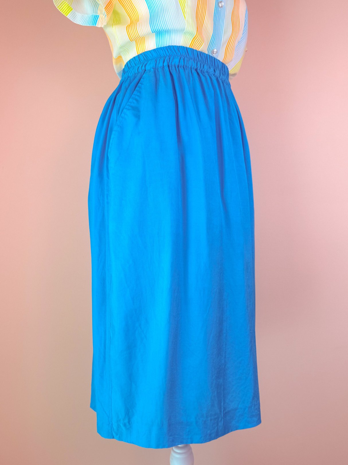 Turquoise blue women's skirt from the 1980s featuring an elastic waistband, displayed on a mannequin against a pink background.