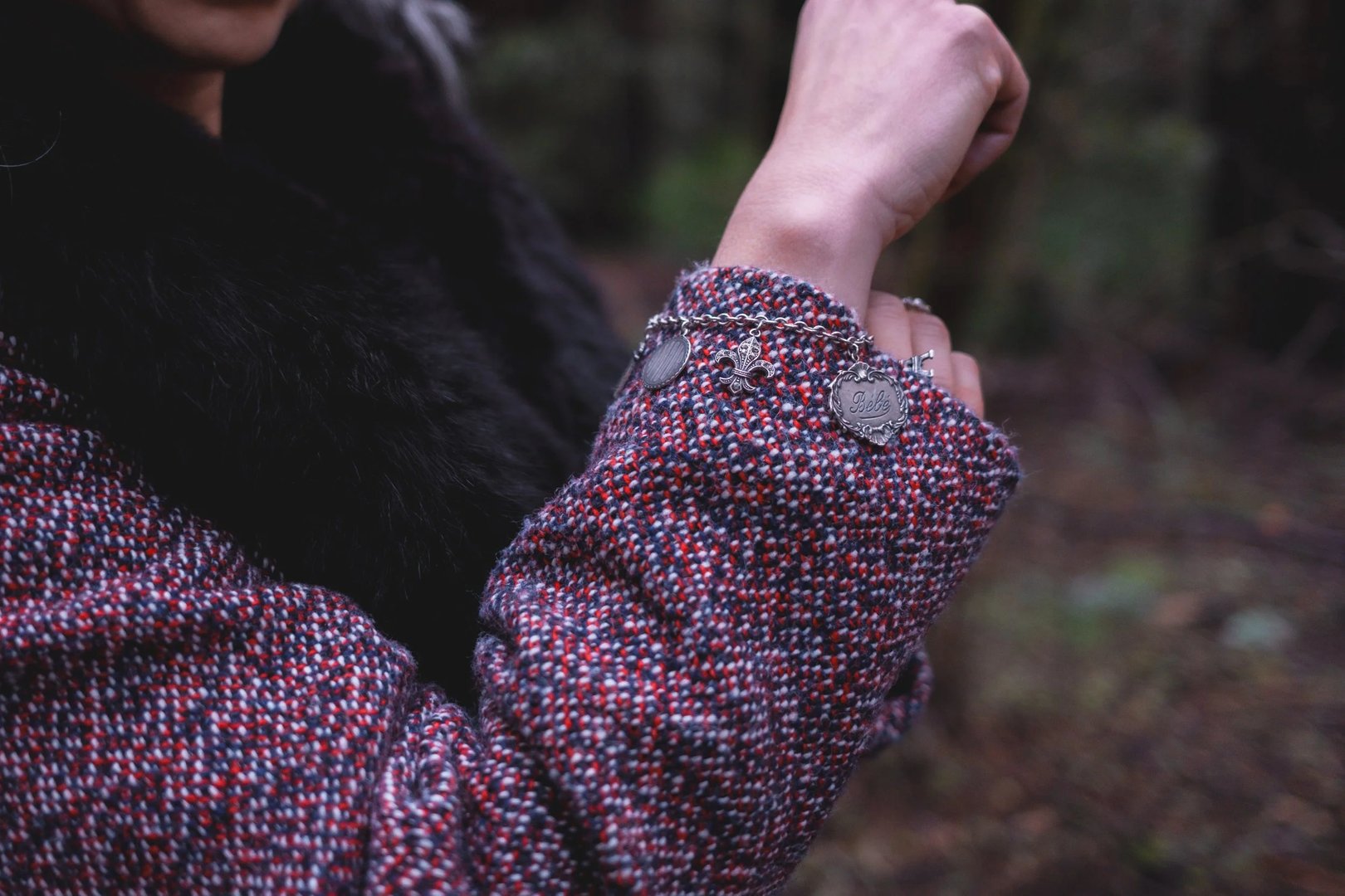 A close-up of a person's wrist adorned with a silver bracelet, wearing a textured, multicolored coat in a forest setting.