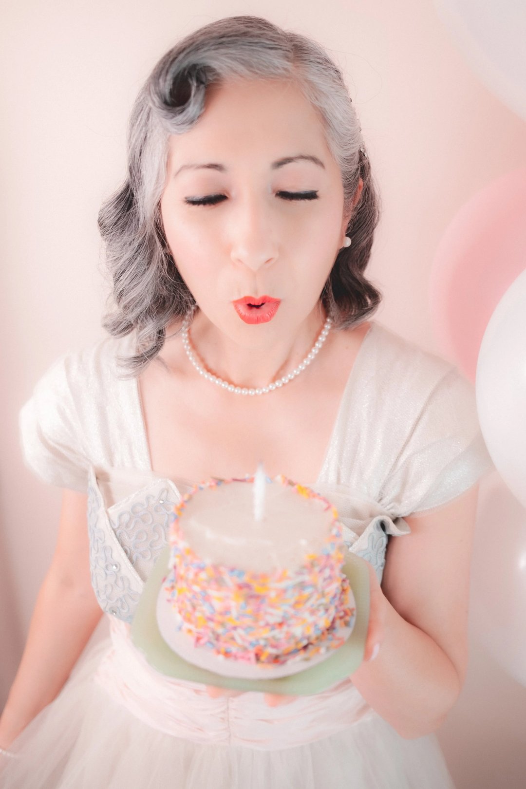 A woman with gray hair blows out a candle on a decorated cake while surrounded by pastel balloons.