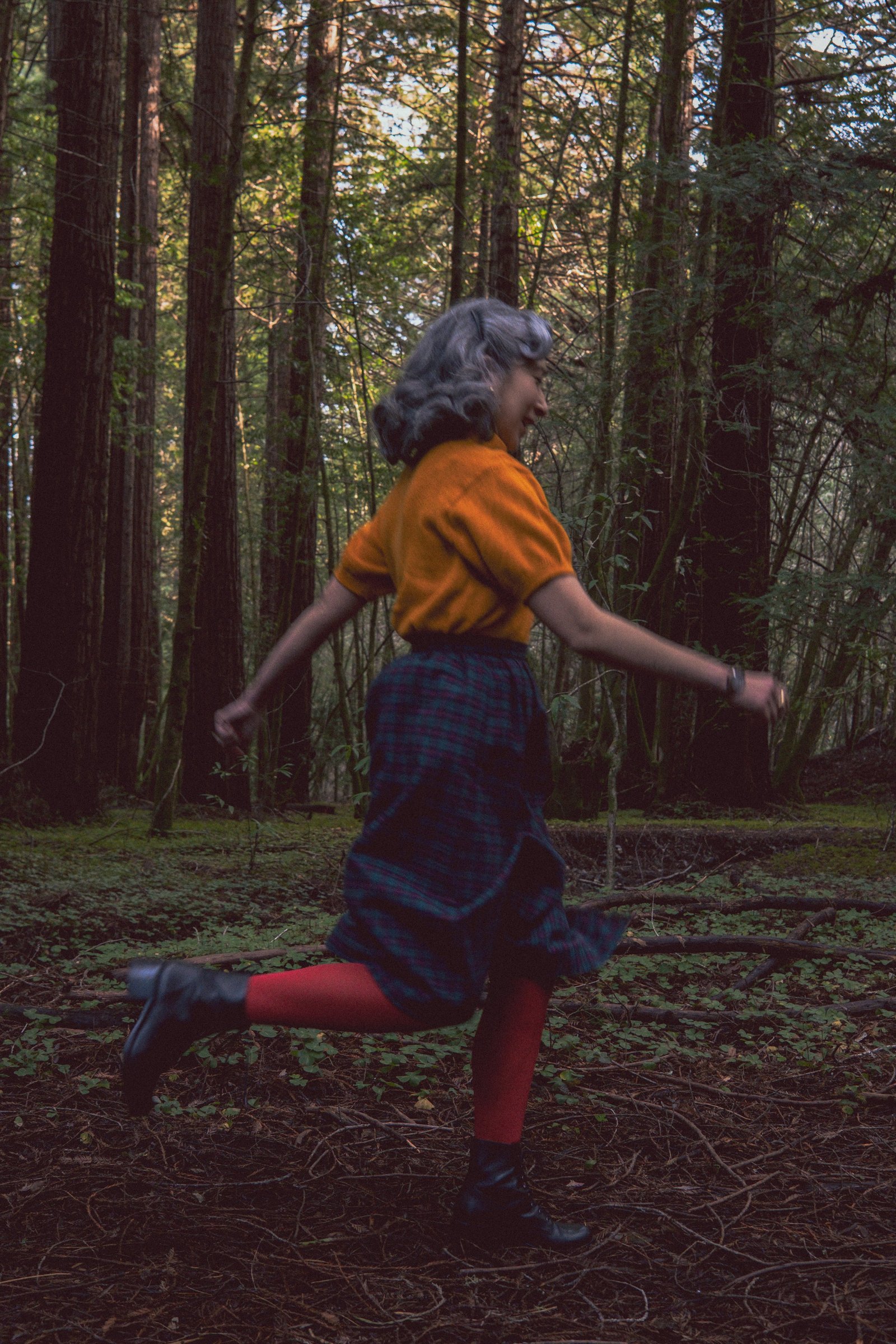 A person with gray hair wearing an orange top, plaid skirt, and red tights is playfully running through a forested area.