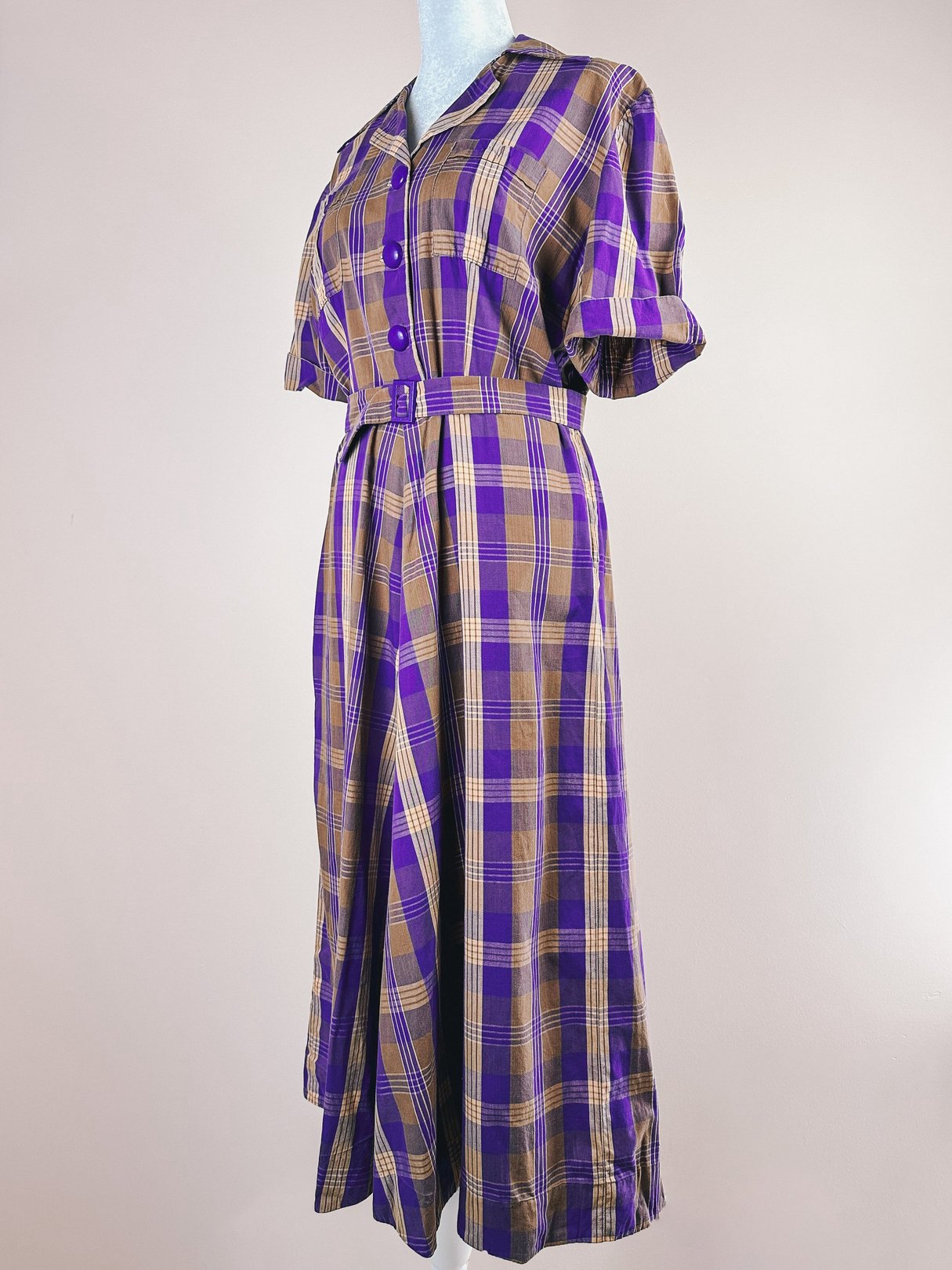 A 1950s women's dress featuring a purple plaid pattern, short sleeves, a button-up front, and a self-tie belt at the waist.
