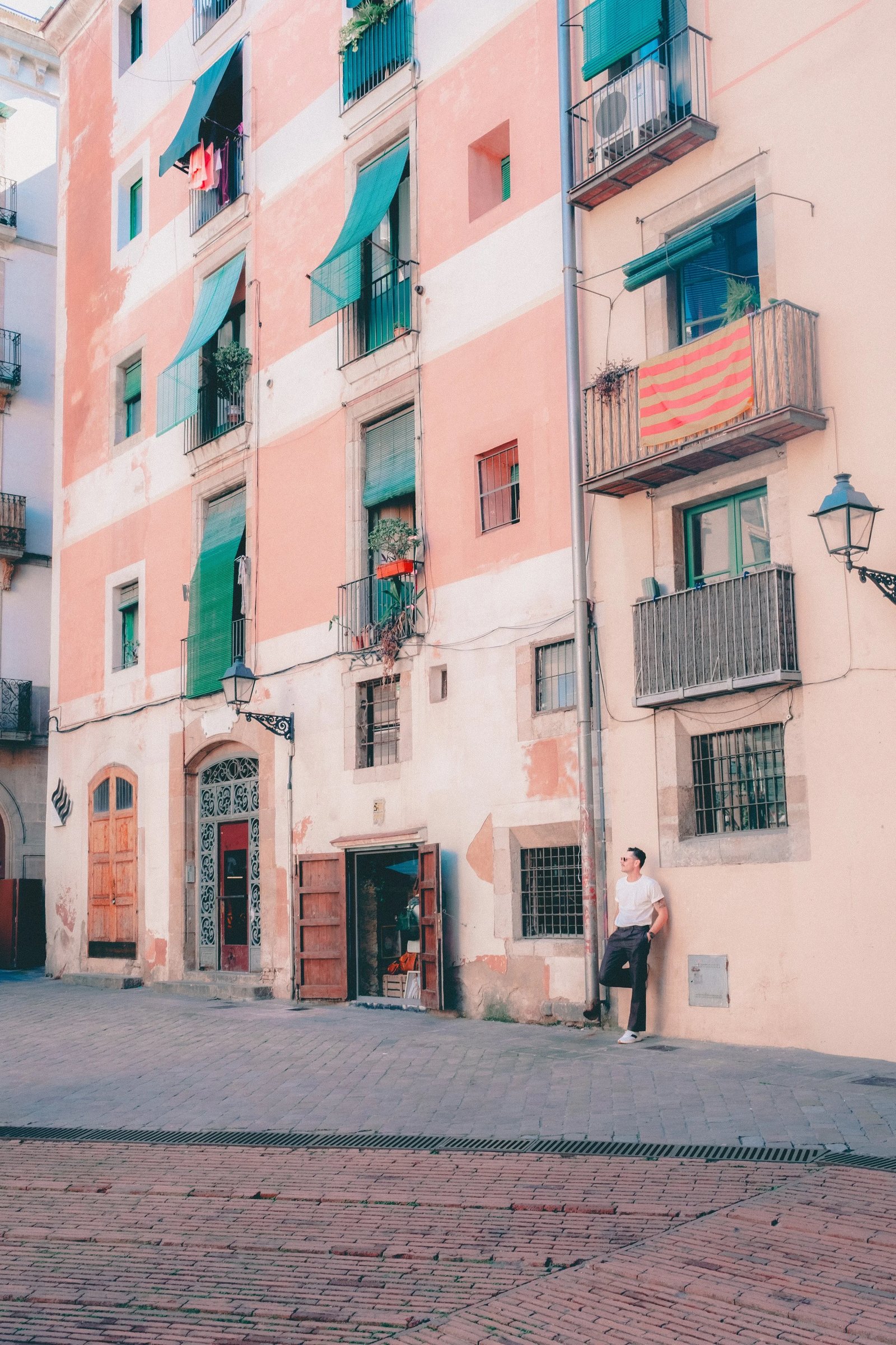 A person leans against a wall in front of a pastel-colored building with balconies and window plants in a narrow street.