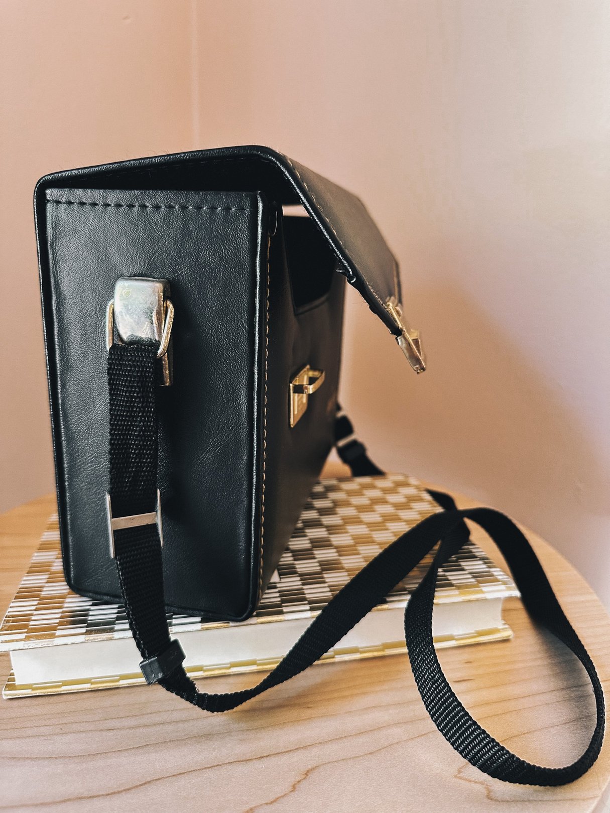 A black leather camera bag with a detachable strap, positioned on top of checkered notebooks, showcasing a vintage 1970s design.