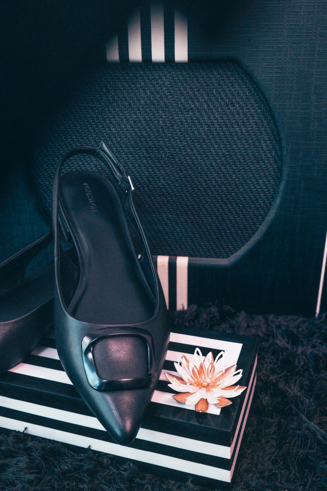 Black slingback shoe with a square buckle displayed on a striped surface alongside a decorative flower.