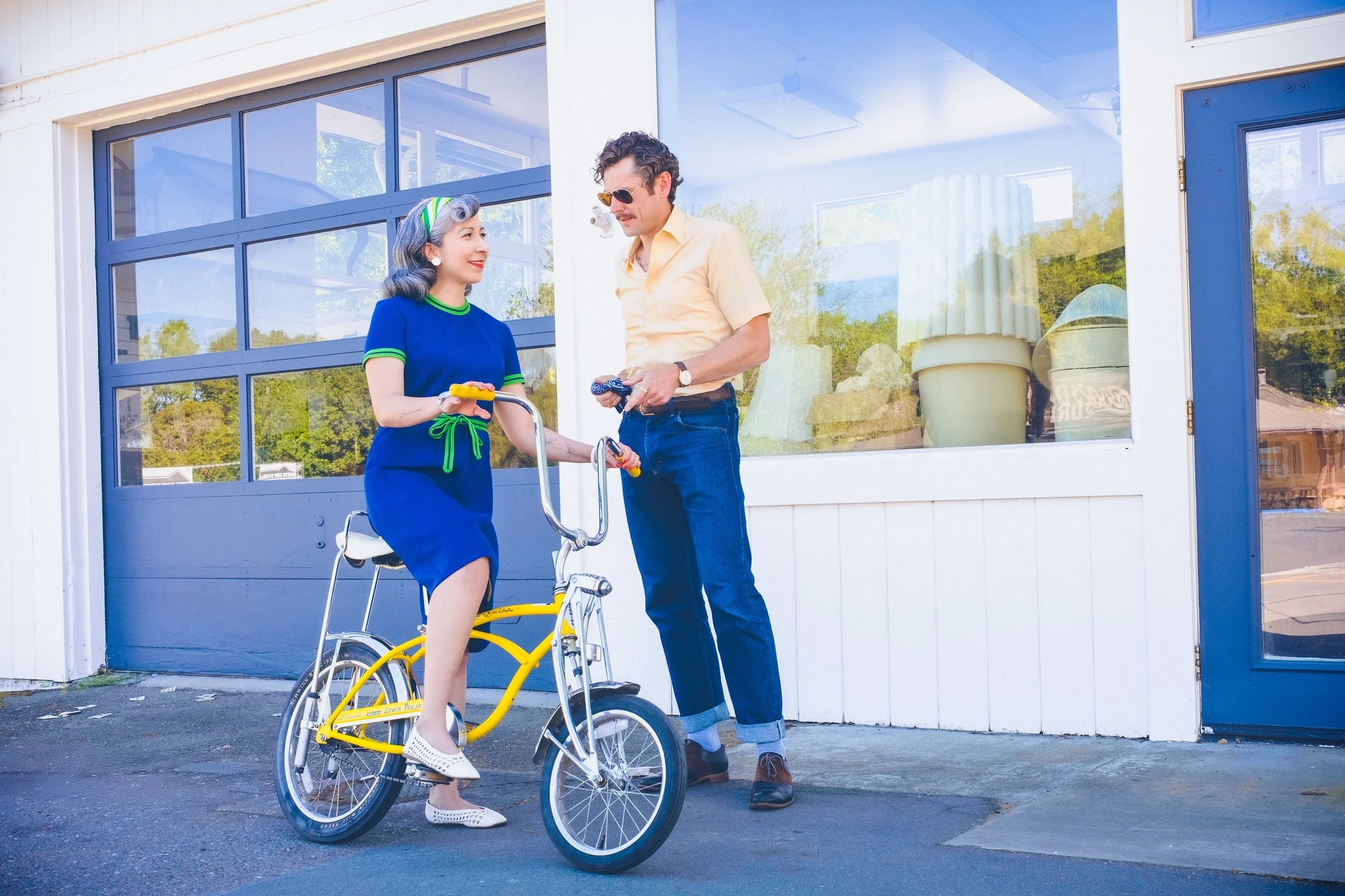 A woman in a blue dress and a man in a yellow shirt stand near a yellow bicycle outside a storefront, both holding drinks.