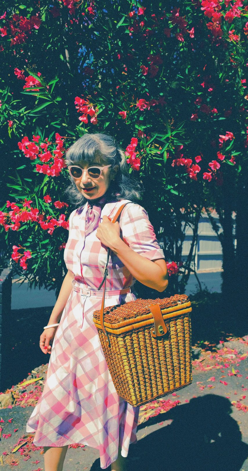 A woman with gray hair, wearing sunglasses and a checkered dress, holds a wicker basket while standing in front of pink foliage.