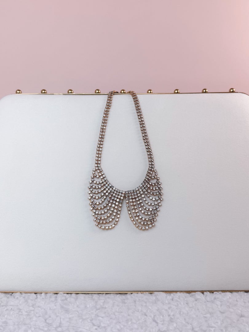 A gold 1970s collar necklace featuring a sparkling design with multiple strands of rhinestones, set against a soft pink background.
