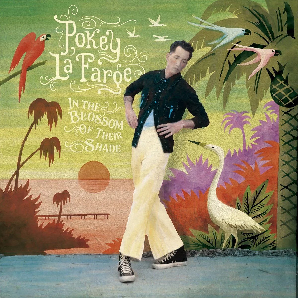 A man in a black button-up shirt and light-colored pants poses against a colorful background featuring tropical plants and a crane.