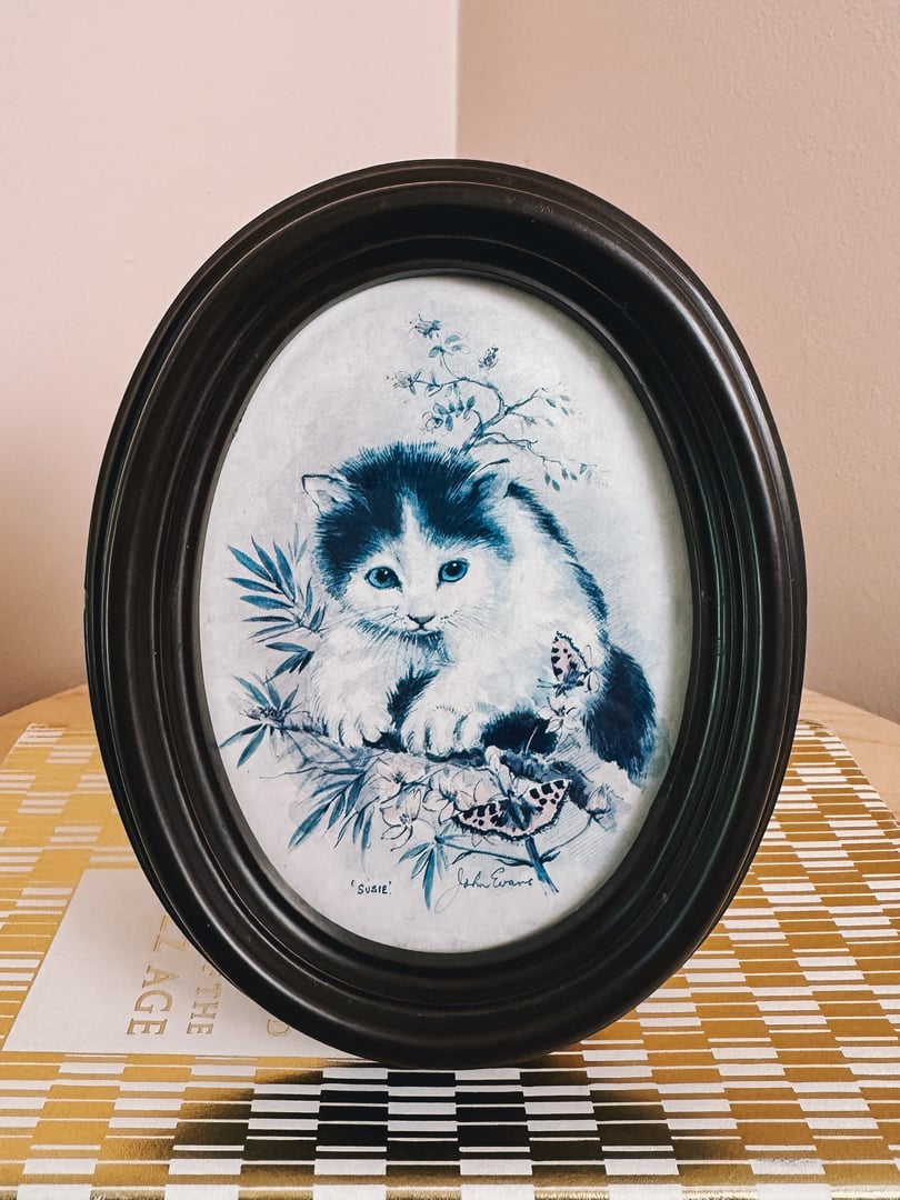 A framed 1970s blue and white portrait of a cat sitting among plants and a butterfly.