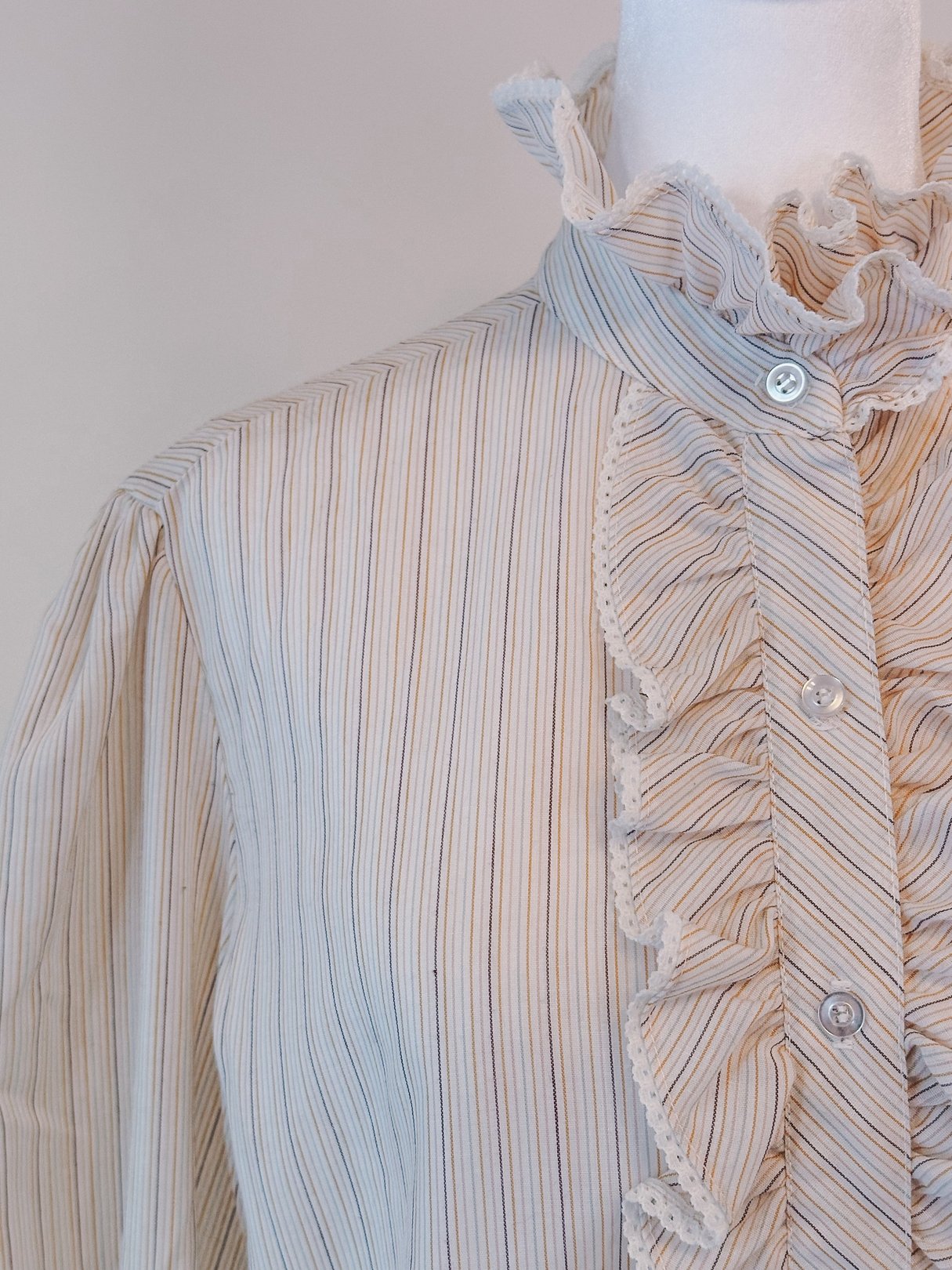 Cream blouse featuring ruffled neck, intricate pink, orange, and blue stripes, and button closure, styled for women in the 1970s.