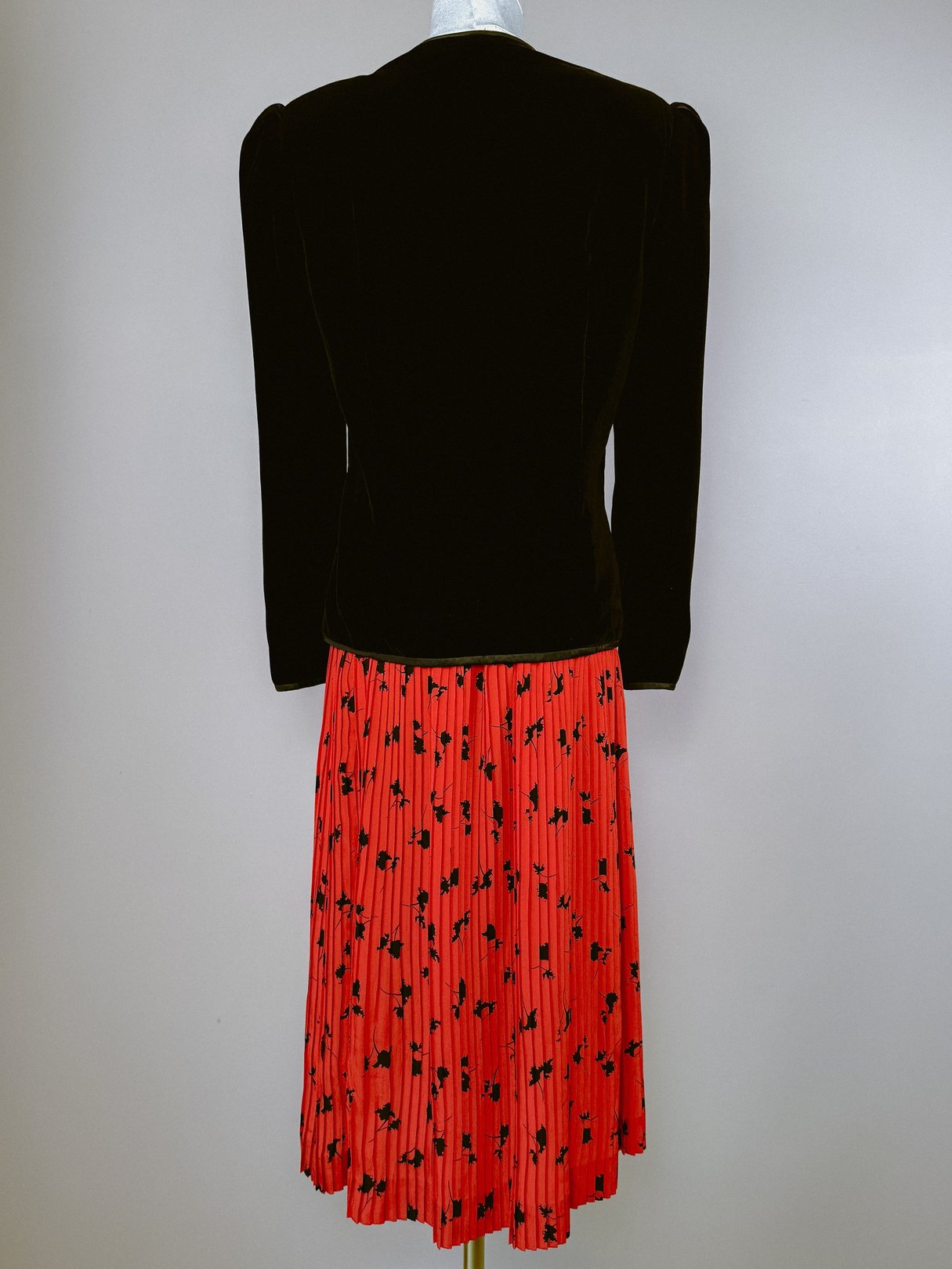 A black velvet jacket with puffed shoulders, styled with a red pleated skirt featuring black floral patterns, representing 1980s women's fashion.