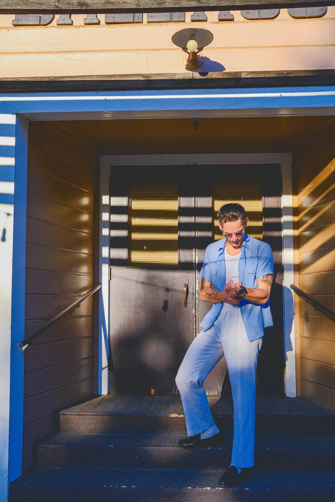 A man in a light blue suit stands on the steps of a building, adjusting his sleeves, with shadows cast by overhead stripes.