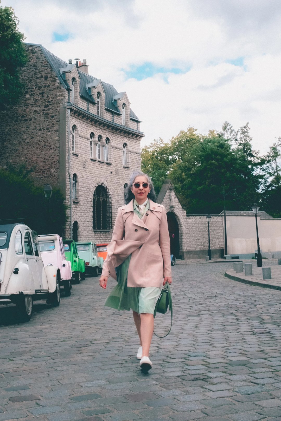 A woman in a light pink trench coat and sunglasses walks along a cobblestone street lined with vintage cars and historic buildings.