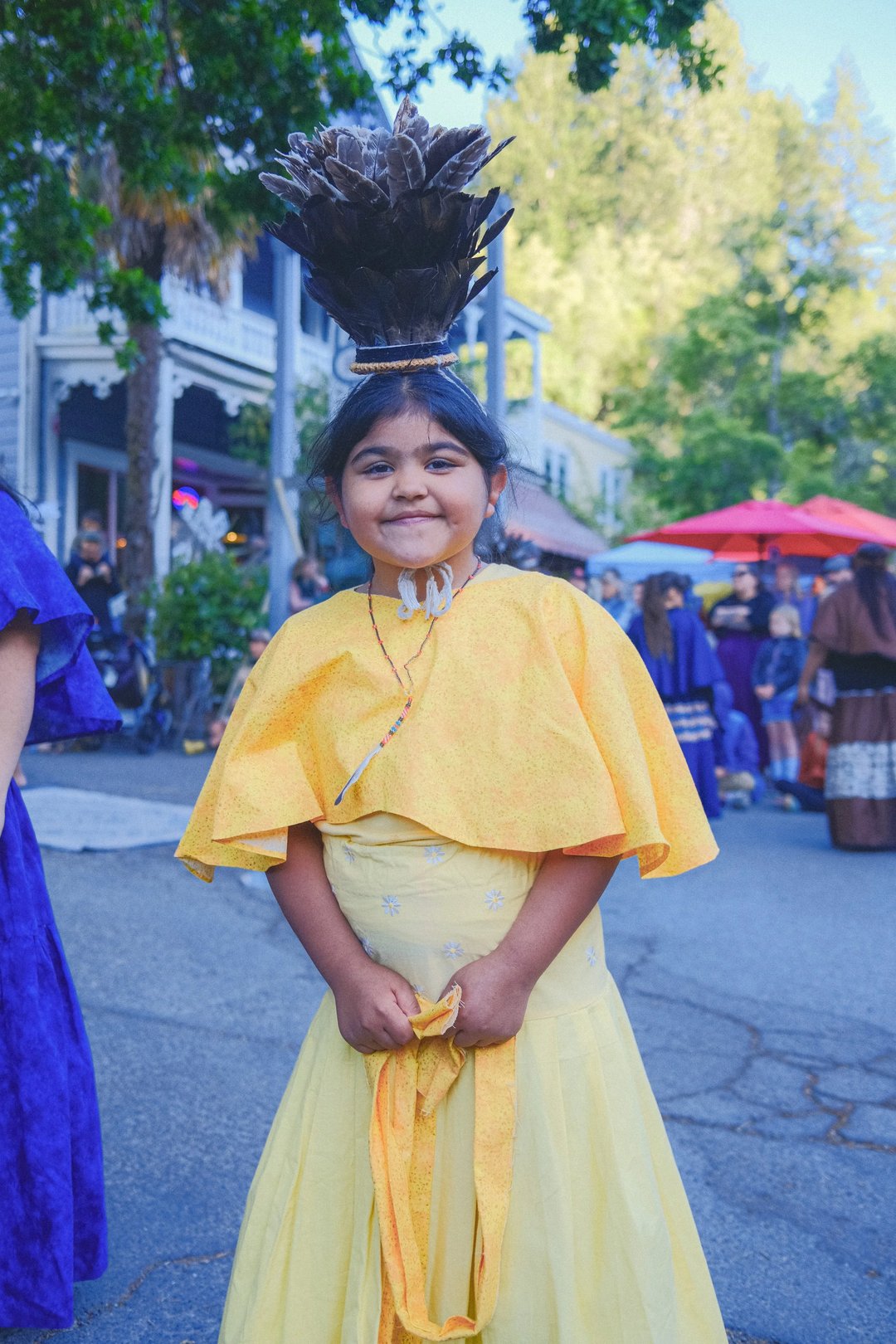 A young girl in a bright yellow dress and matching cape smiles at the camera during a cultural event.