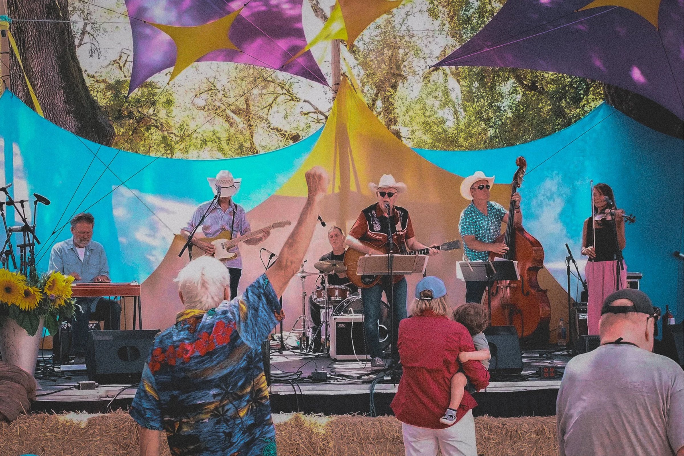 A band performs on stage at an outdoor event with colorful fabric canopies while an audience member raises a fist in celebration.