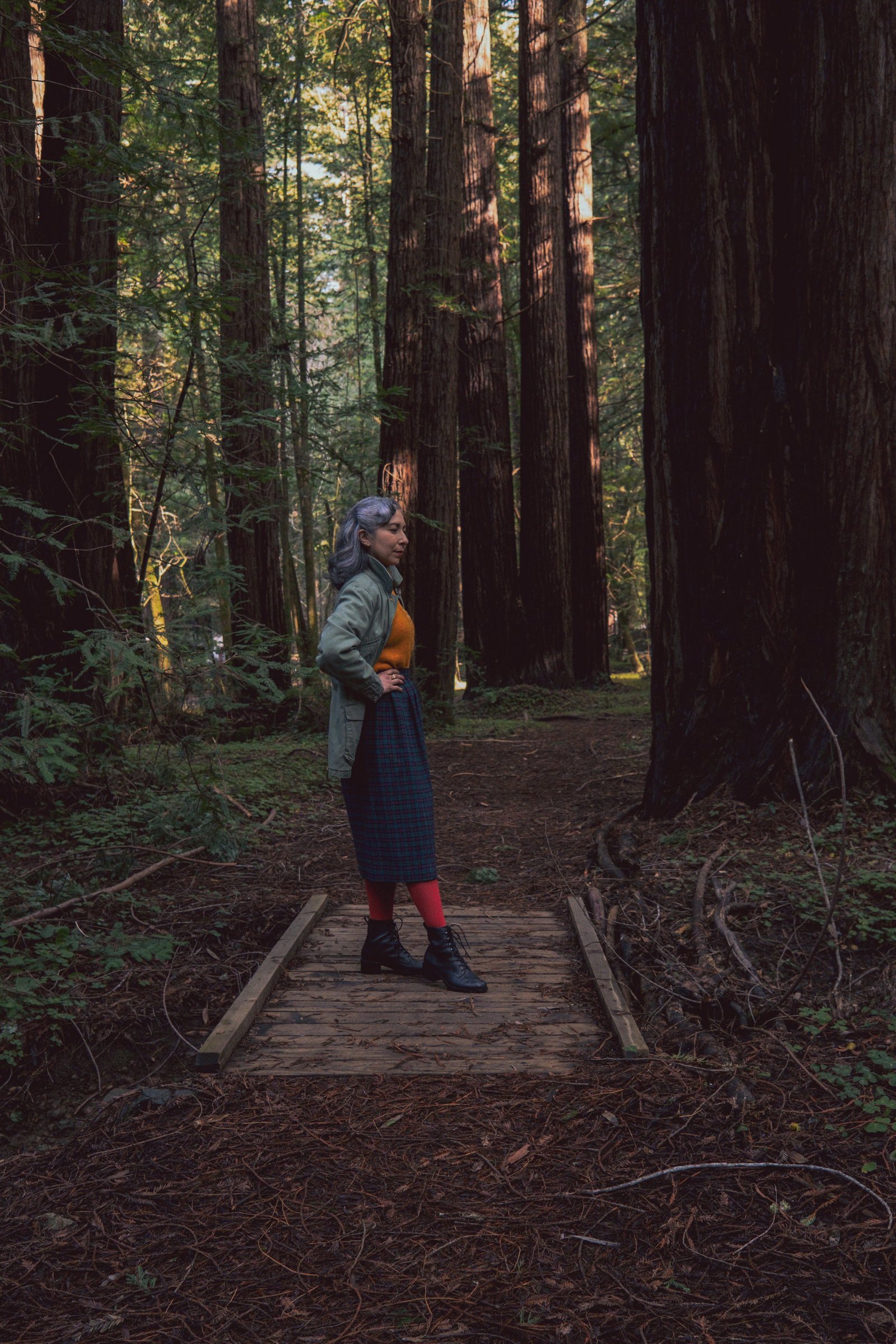 A woman stands on a wooden path in a forest of tall redwood trees, wearing a green jacket, orange top, plaid skirt, and black boots.