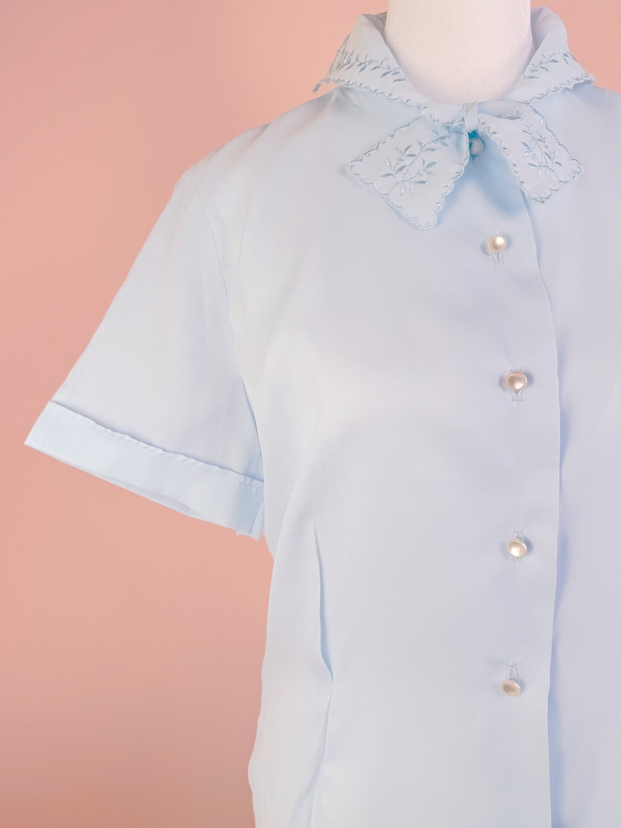 Powder blue blouse for women with short sleeves, a collar featuring embroidered details, and pearl-button closures, reminiscent of 1950s fashion.