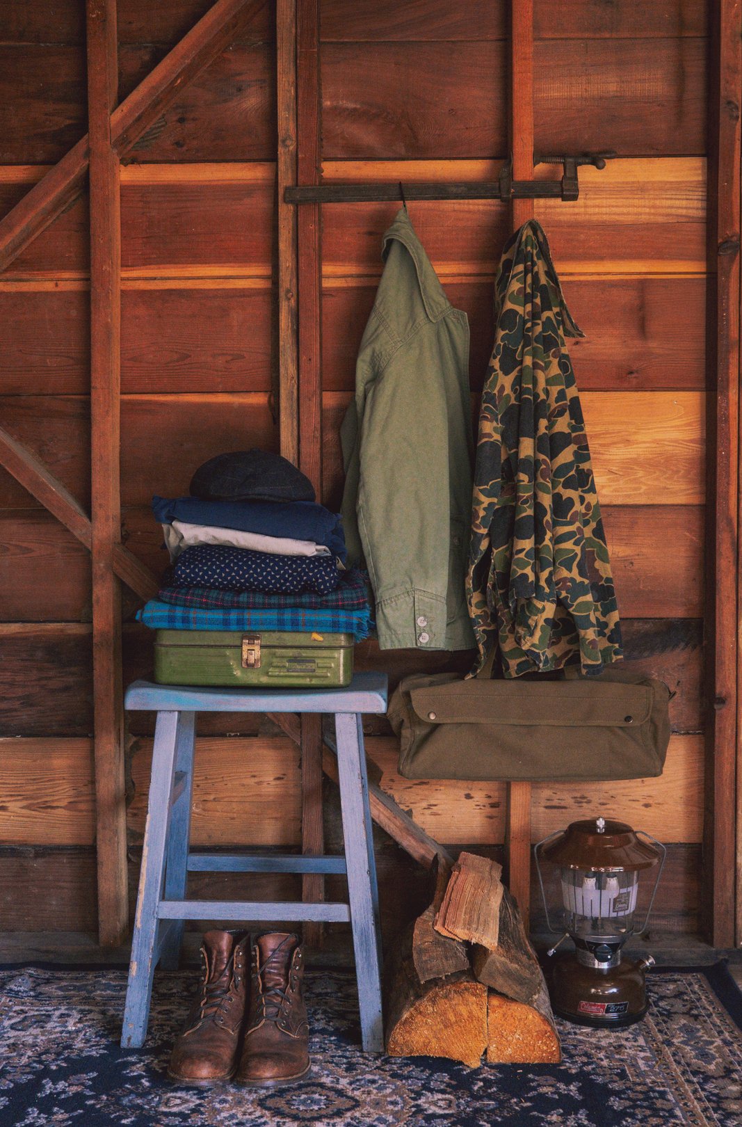 A rustic wooden shed interior featuring a purple stool with stacked clothing and three hanging jackets.